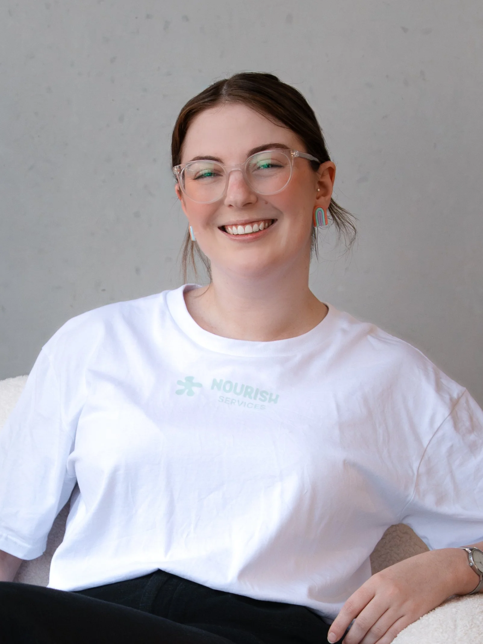 A woman with brown hair, wearing glasses, earrings, and a white t-shirt, smiling and sitting against a plain wall.