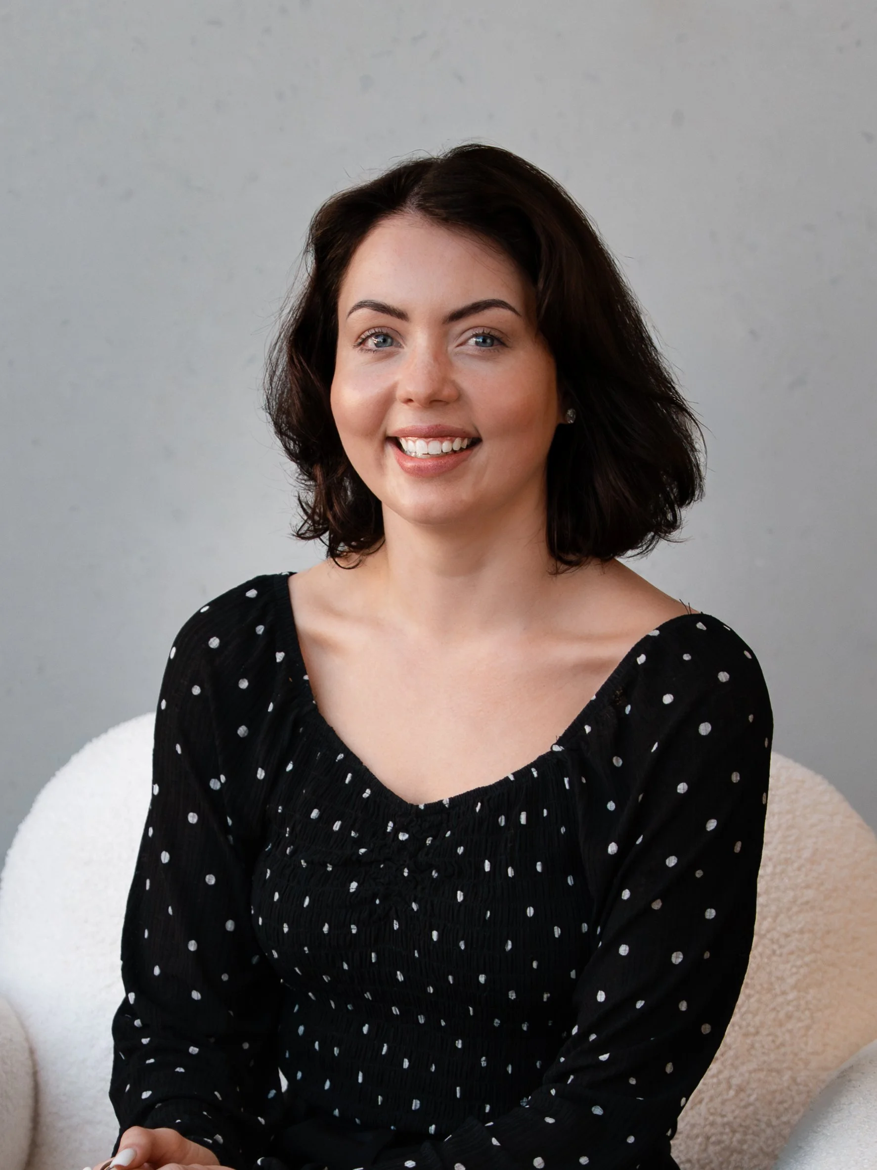 A woman with short dark hair, blue eyes, and fair skin sitting on a white chair against a plain gray background, smiling at the camera, wearing a black dress with white polka dots.