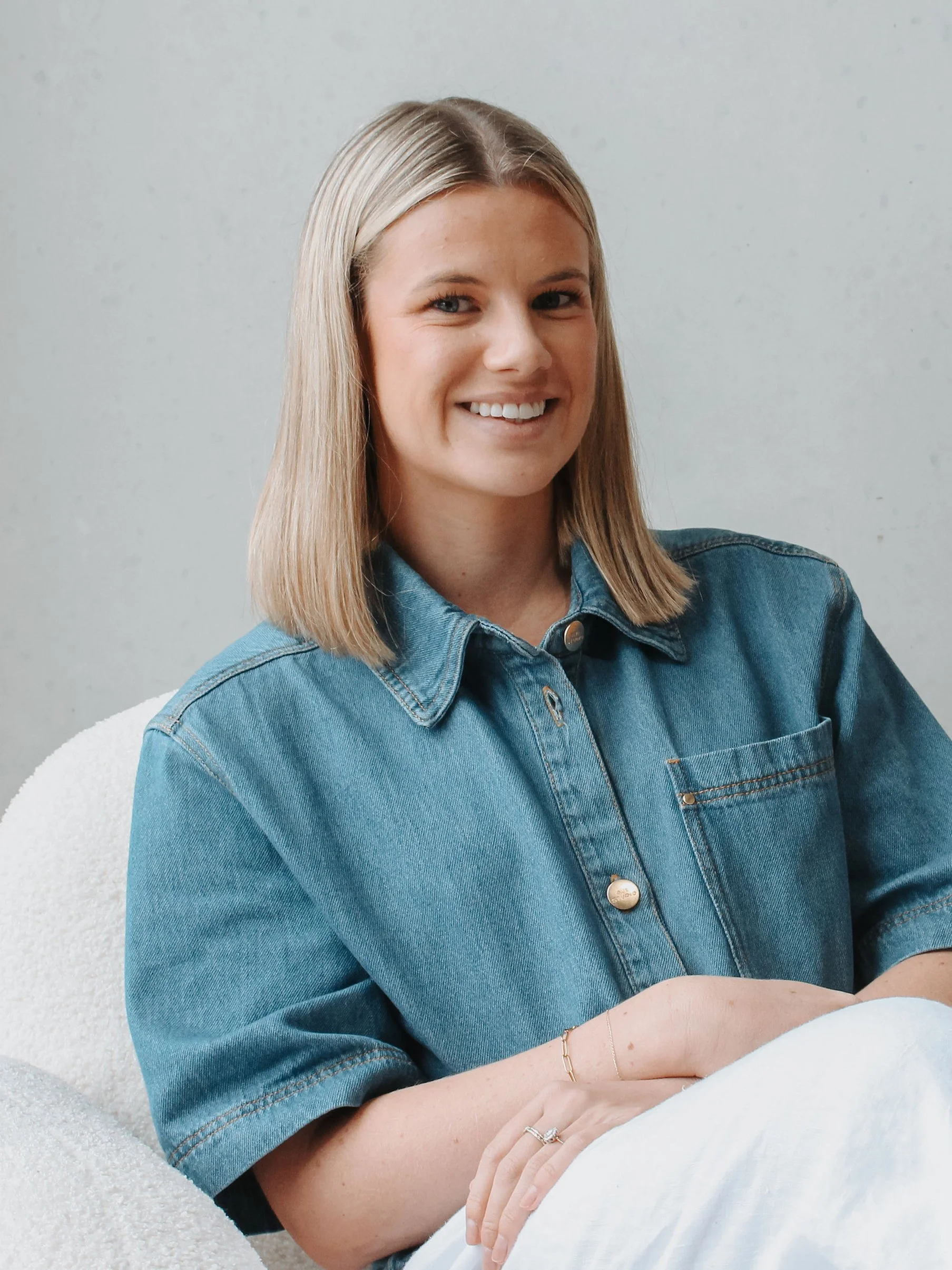 A woman with blonde hair wearing a denim jacket and a bracelet, smiling while sitting on a white chair against a plain light background.