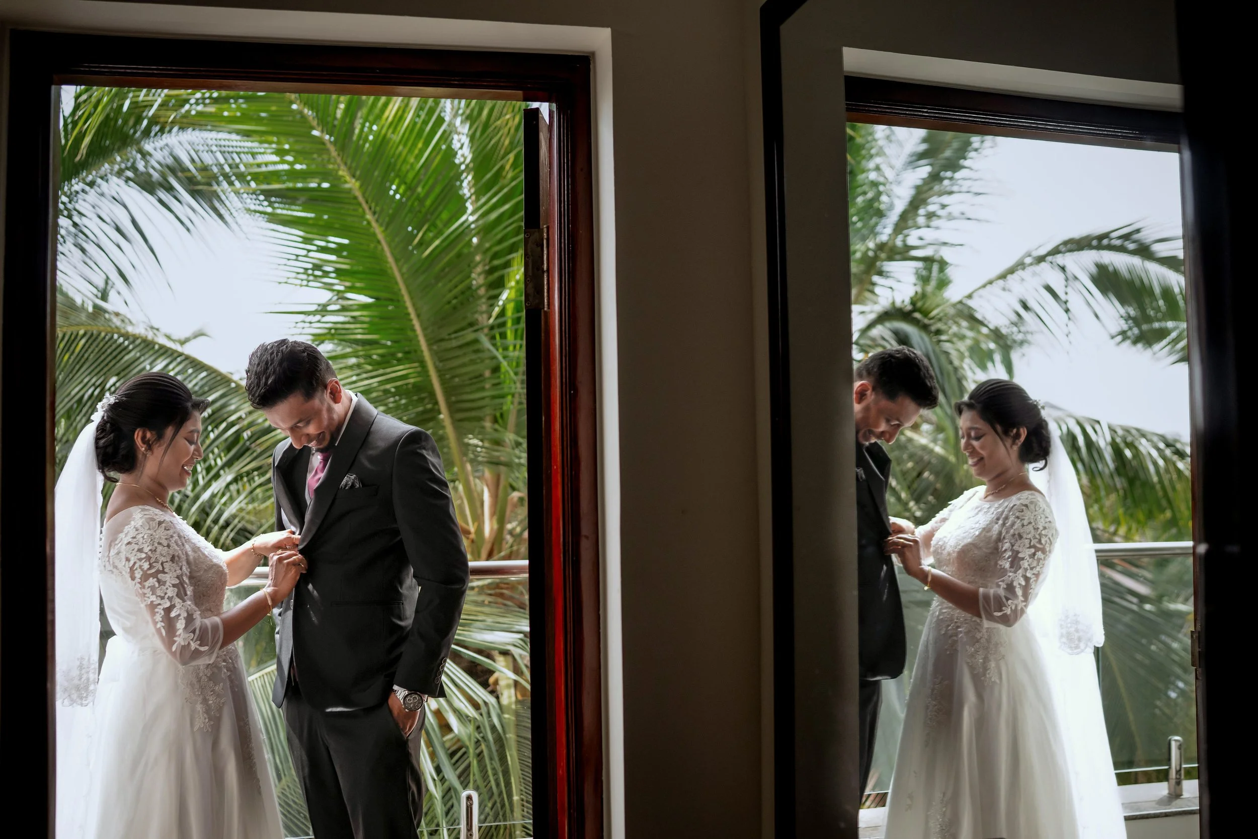 A couple in wedding attire standing on a balcony, with green palm trees in the background. The bride is adjusting the groom's suit, and they both are smiling.