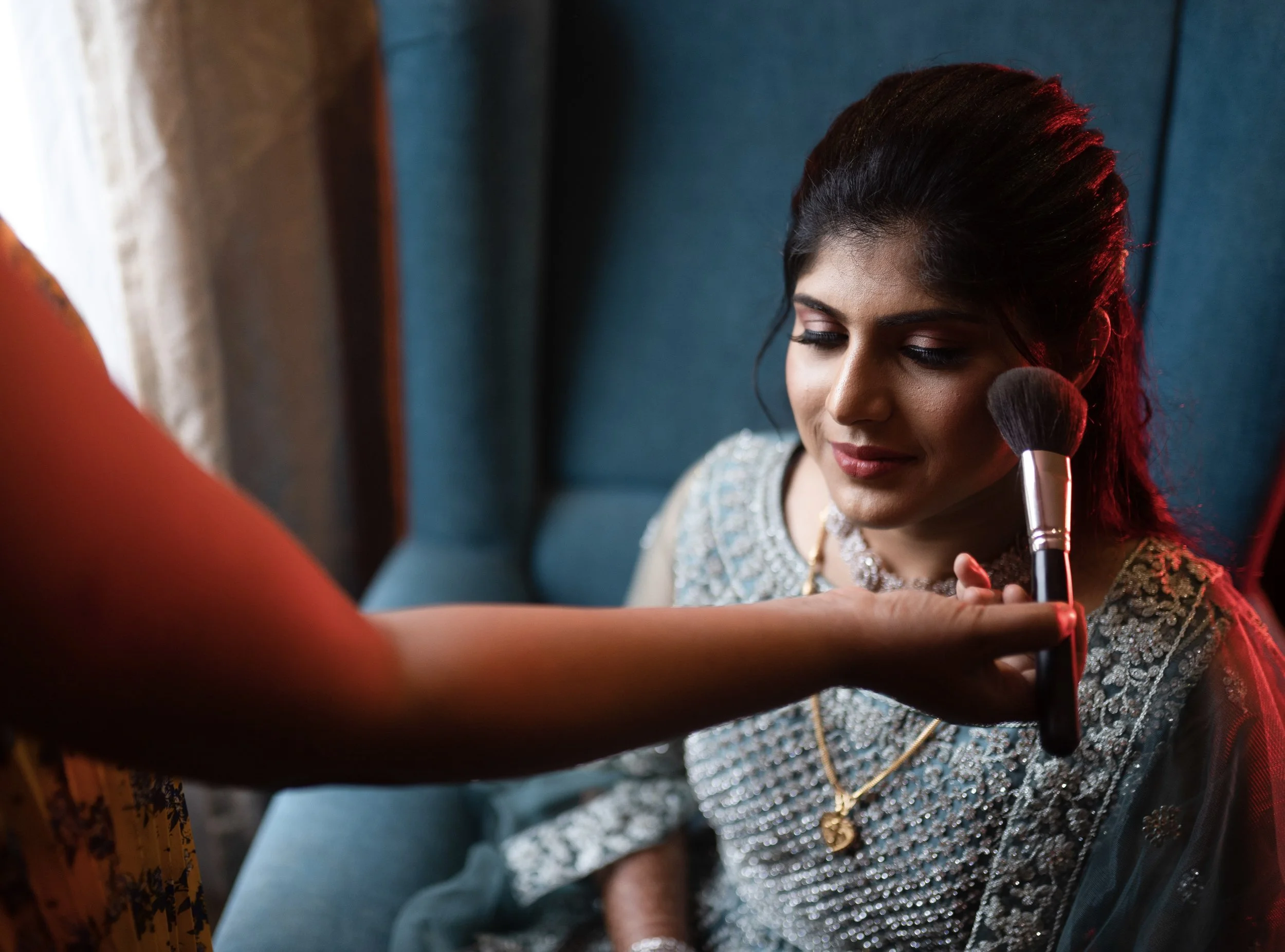 The Bride with dark hair and traditional Indian attire, sitting with her eyes closed as a makeup artist applies makeup with a brush.