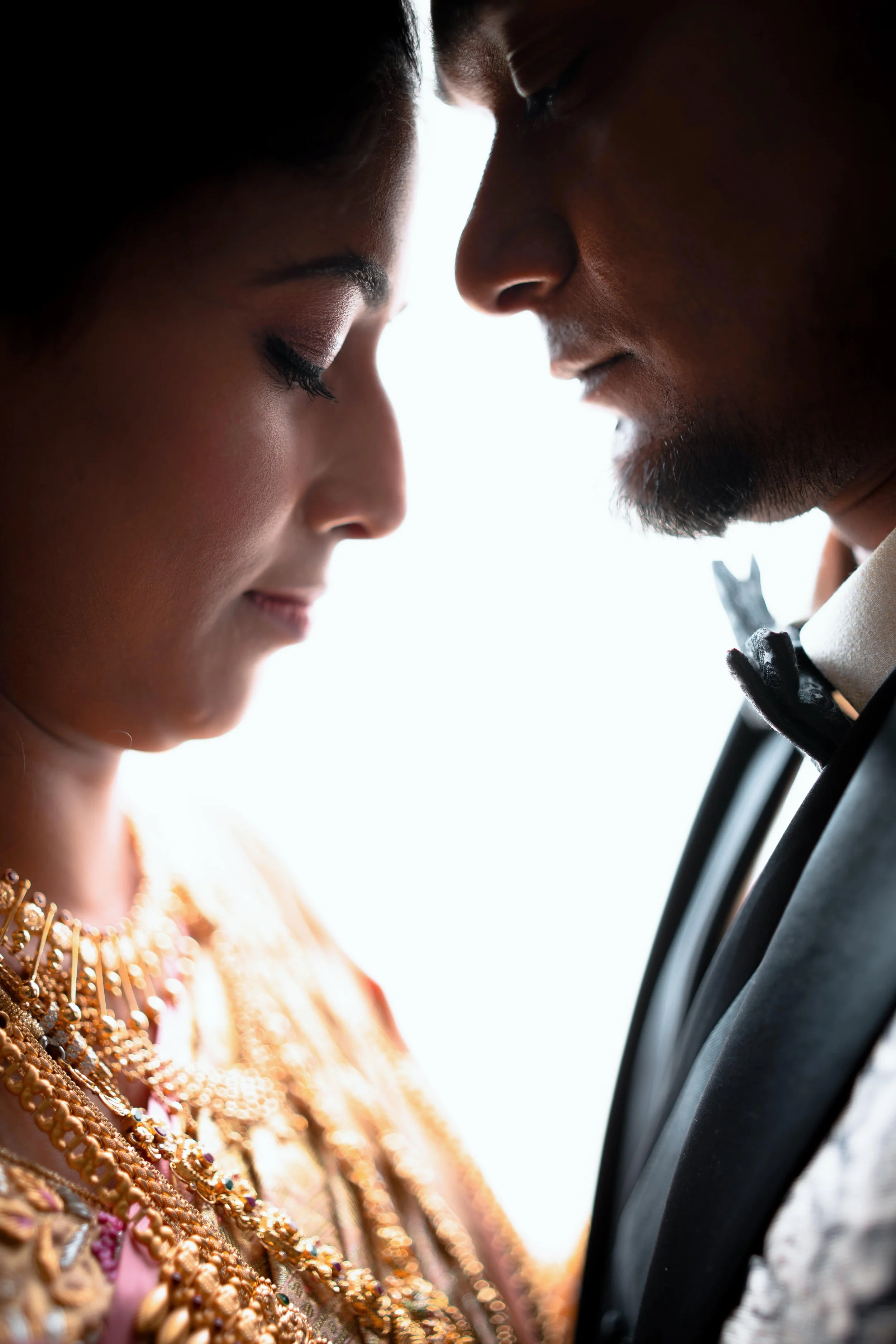 Close-up of a couple with foreheads touching, eyes closed, in a romantic pose. The woman is dressed in traditional Indian attire with jewelry, and the man is wearing a tuxedo with a bow tie.