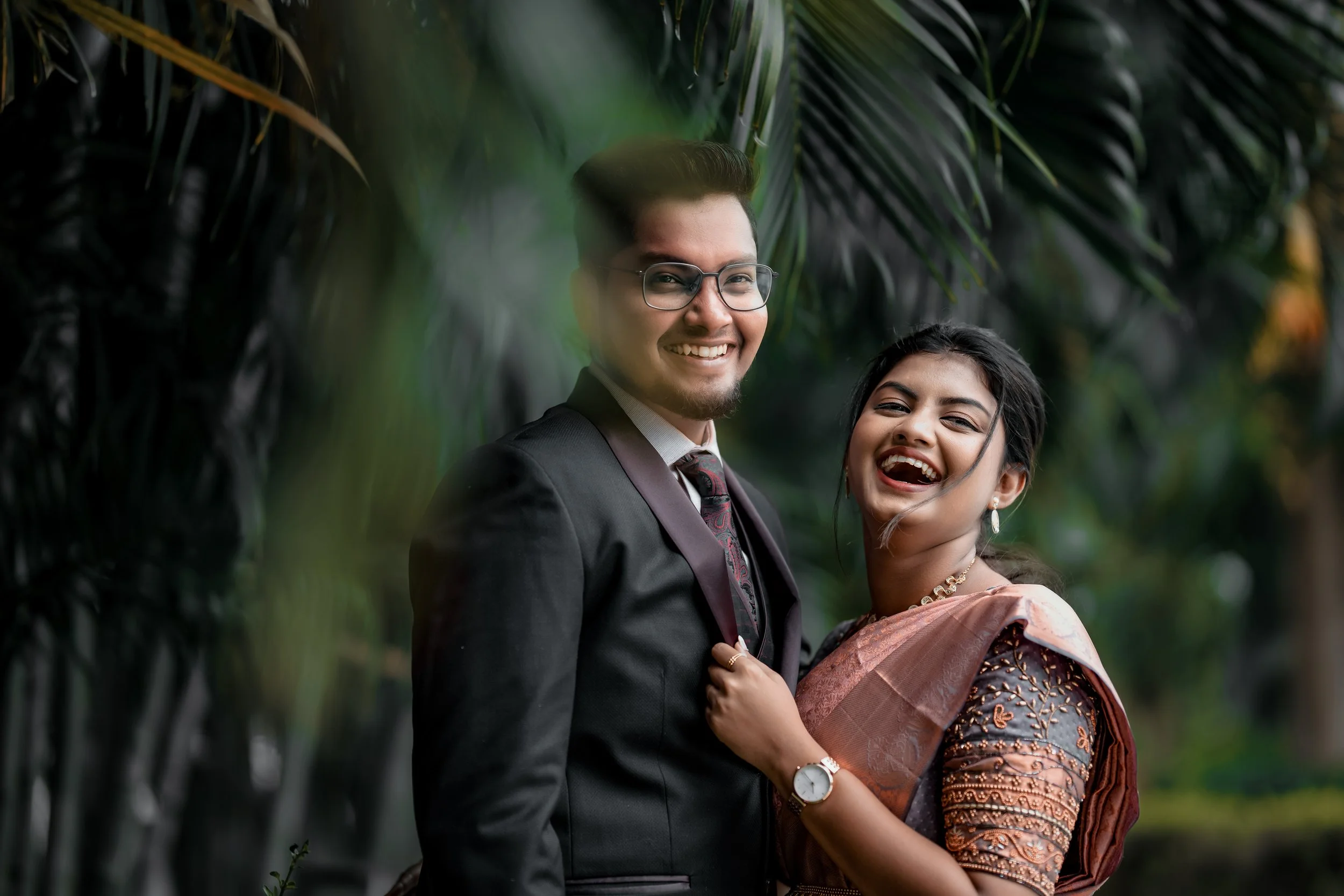 A smiling couple standing close together outdoors, surrounded by lush greenery. The man is wearing glasses, a dark suit, and a tie, while the woman is dressed in traditional attire with jewelry. They look happy and are in a joyful mood.