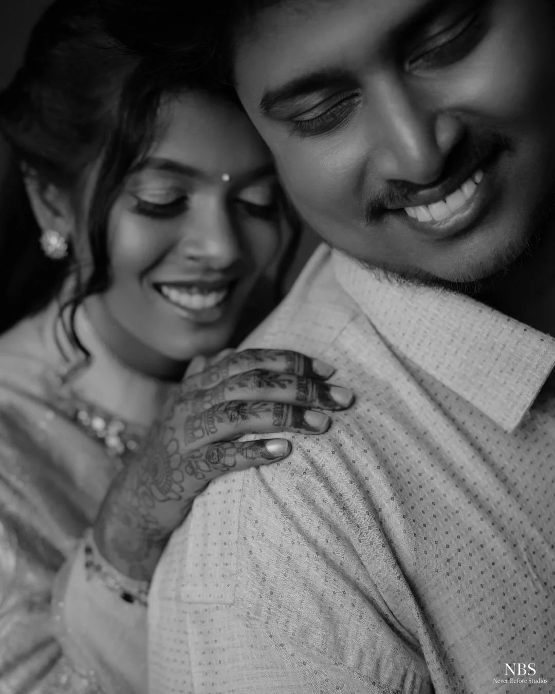 A black and white photo of a smiling couple embracing, with intricate henna designs on the woman's hand resting on the man's shoulder.