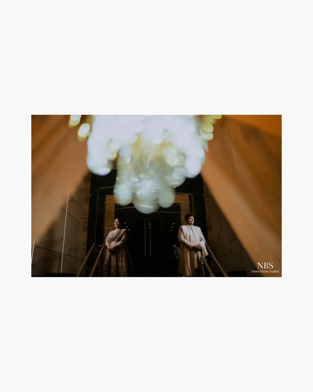 A couple in traditional attire standing at the entrance of a building, viewed from below through a chandelier and surrounded by warm lighting.