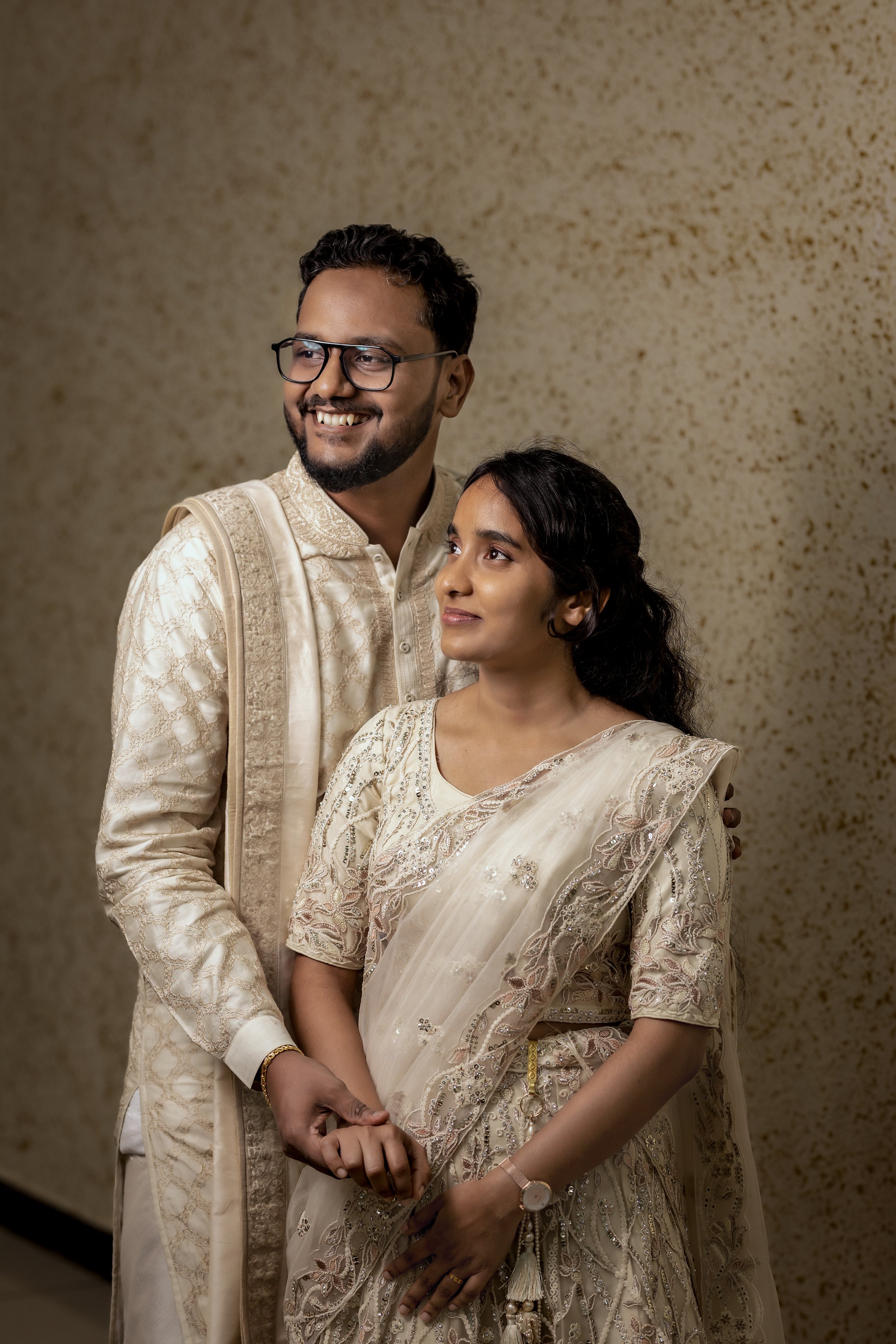 A man and woman in traditional Indian attire standing close together, smiling, with a textured beige background.