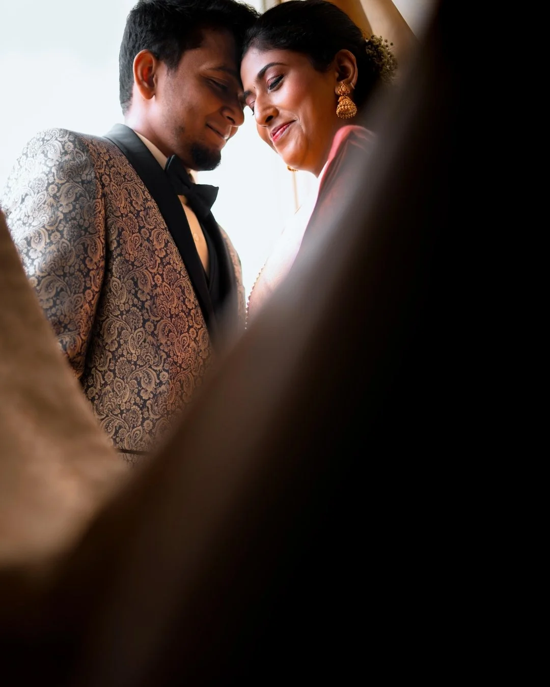 A couple dressed in formal attire sharing an intimate moment, with heads touching and eyes closed, smiling softly.
