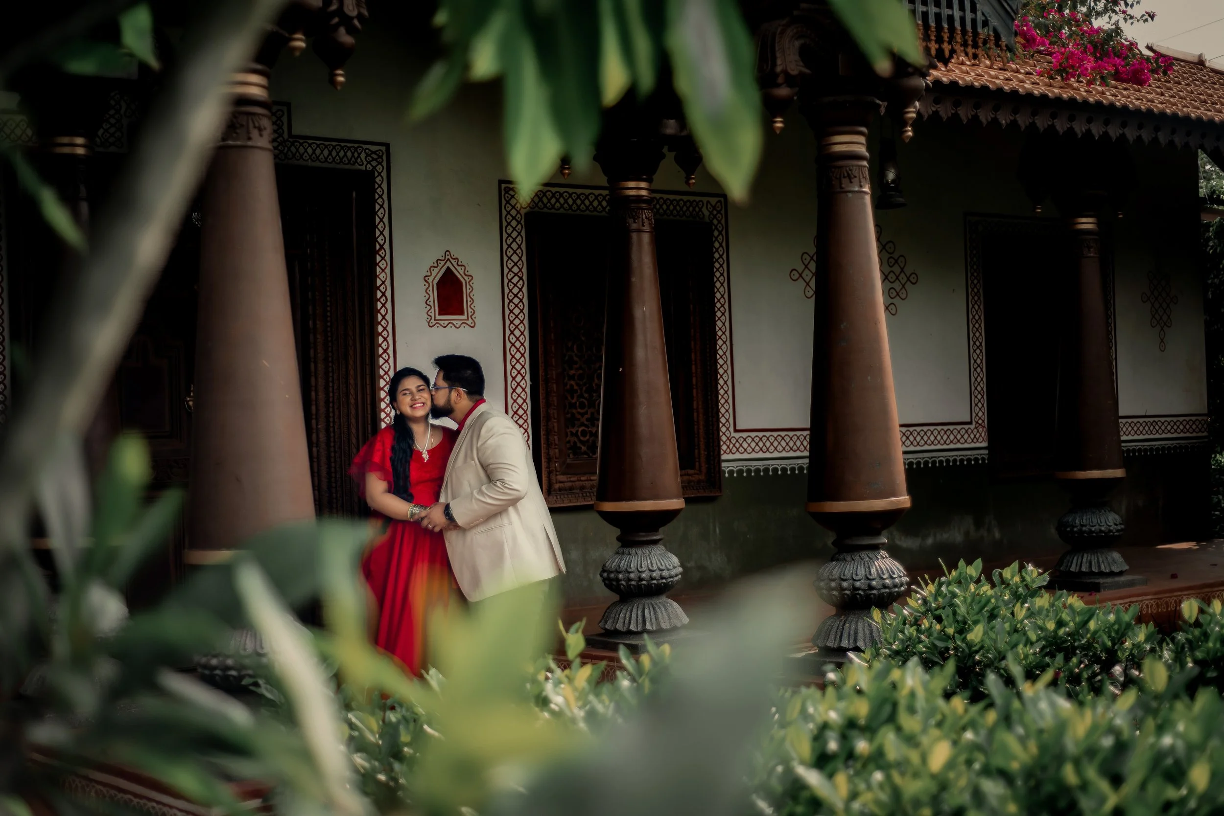 A couple dressed in traditional Indian attire standing close together and smiling, with the man kissing the woman on the cheek, in front of a decorated and traditional building with large wooden columns and ornate window frames, partially obscured by