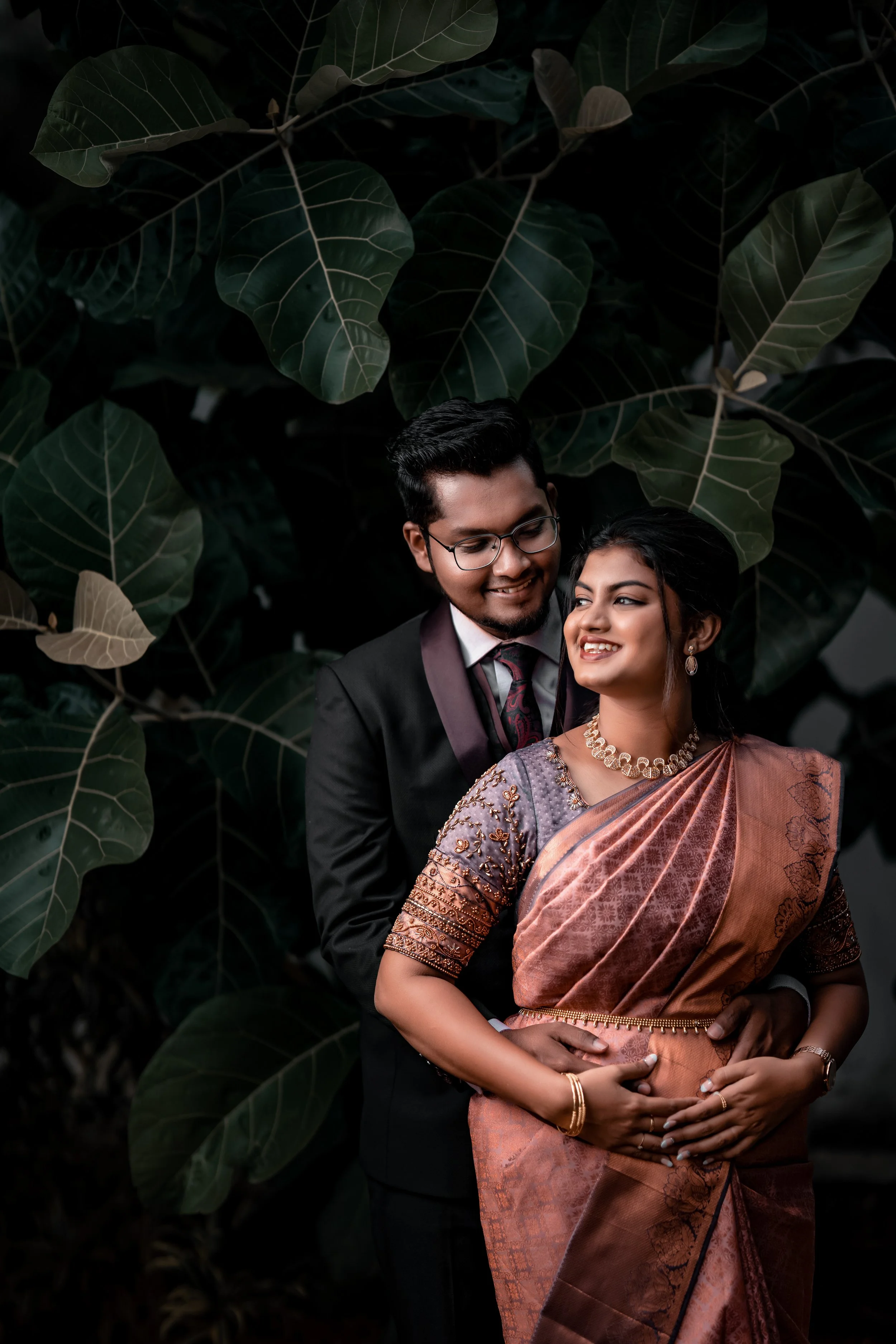 A happy couple dressed in traditional Indian attire, standing closely together with the man embracing the woman from behind, against a backdrop of large green leaves.