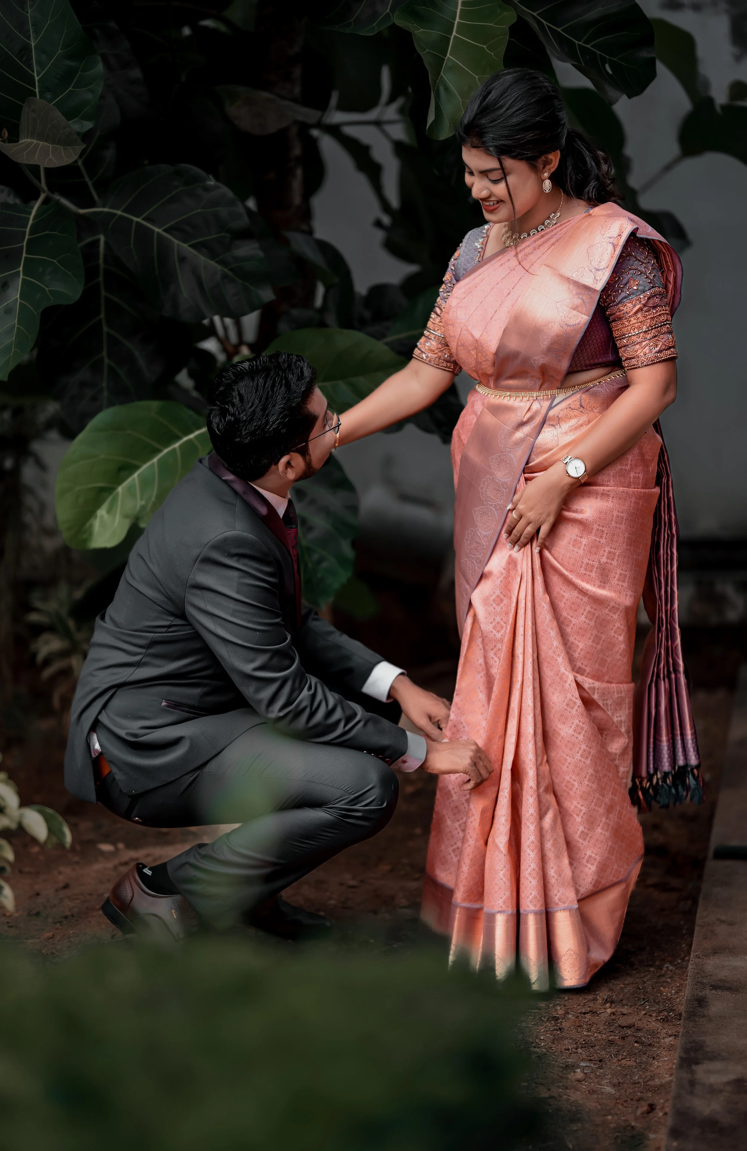 A man in a suit kneeling and adjusting the hem of a woman's sari, who is dressed in a traditional pink and brown sari, in an outdoor garden setting with large green leaves.