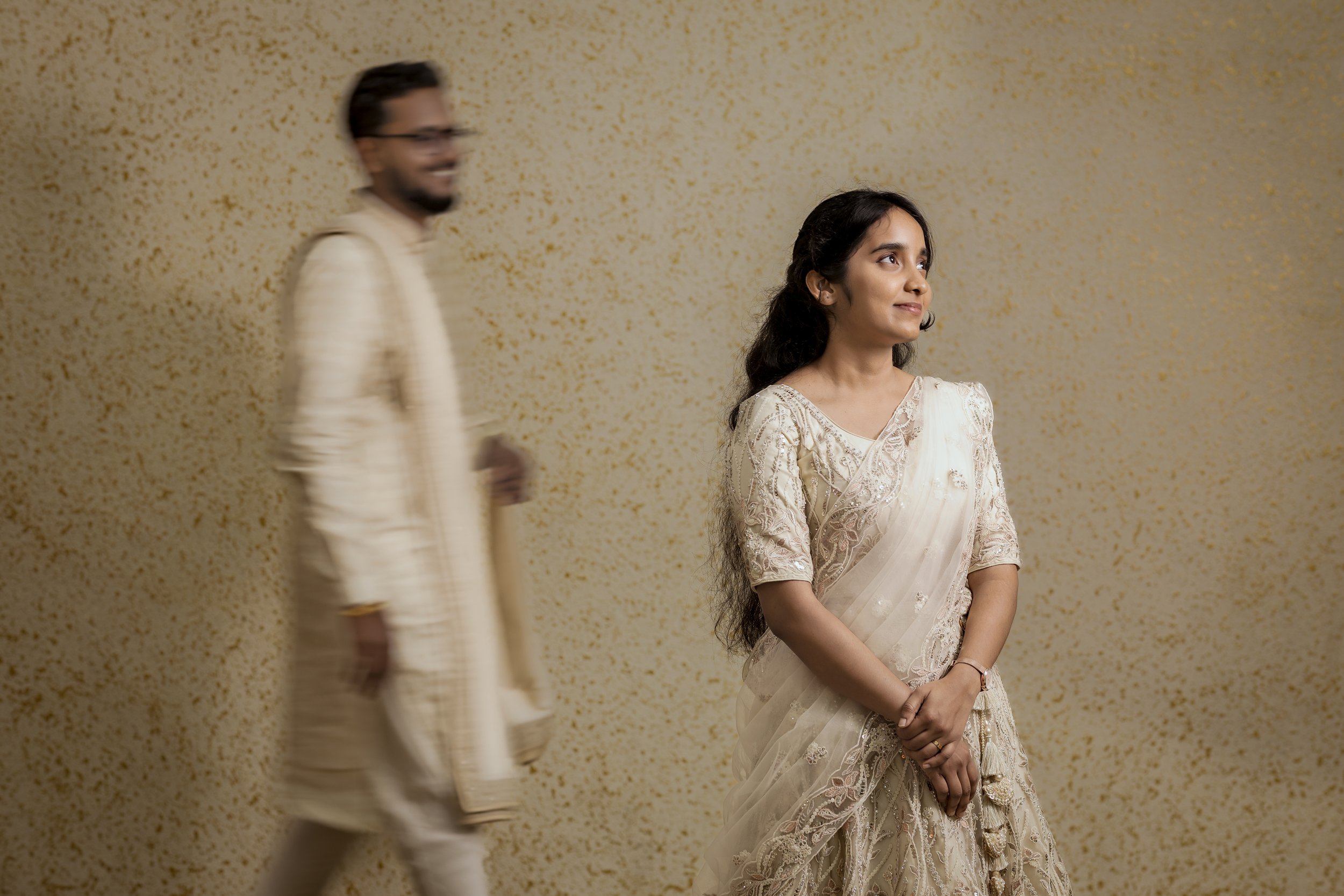 A woman in a cream-colored traditional Indian outfit standing in front of a beige and gold speckled background, with a man in cream-colored attire walking past her in the background, which is slightly blurred.