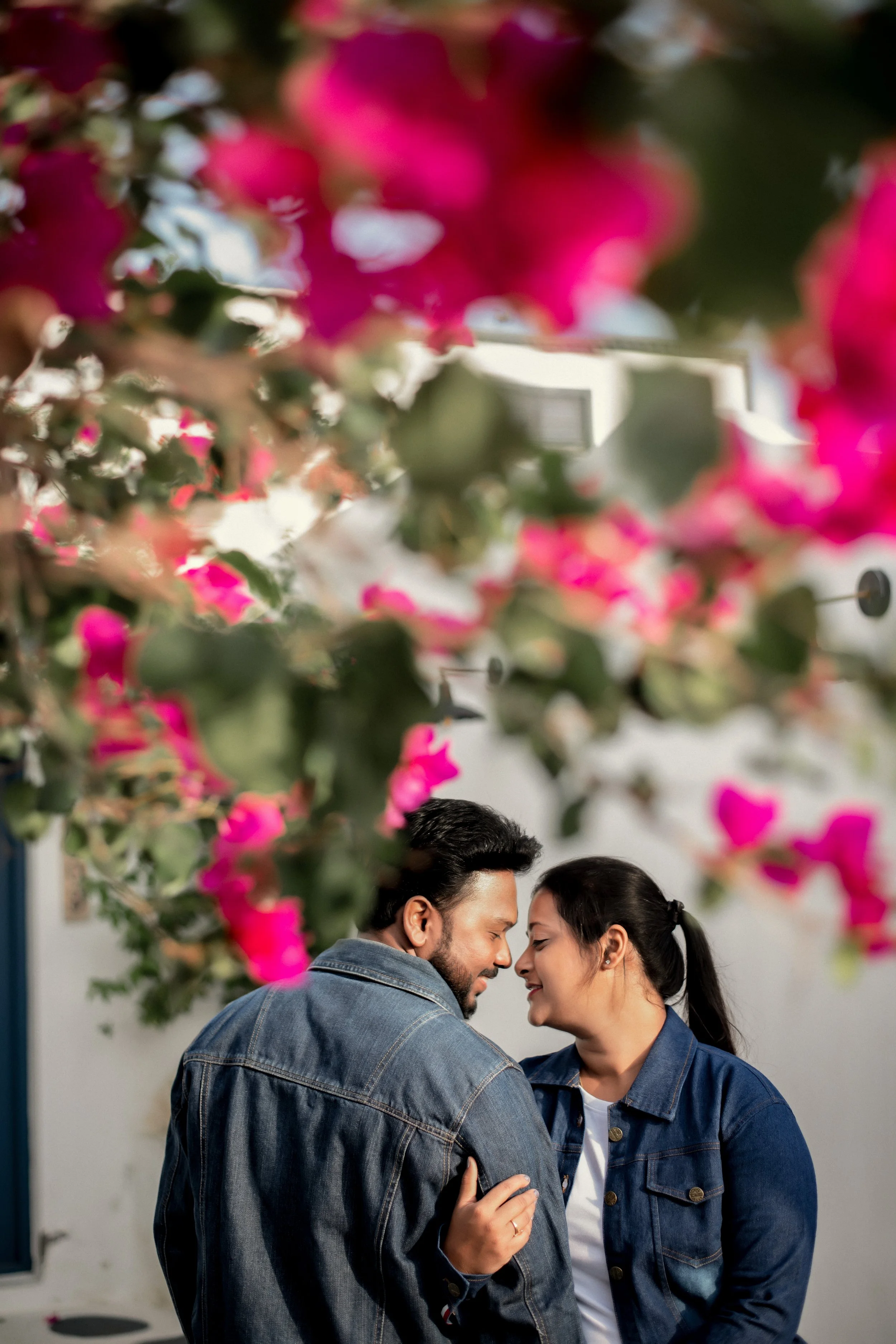 Bride and groom are close together, smiling and touching foreheads, surrounded by pink flowers in the foreground.