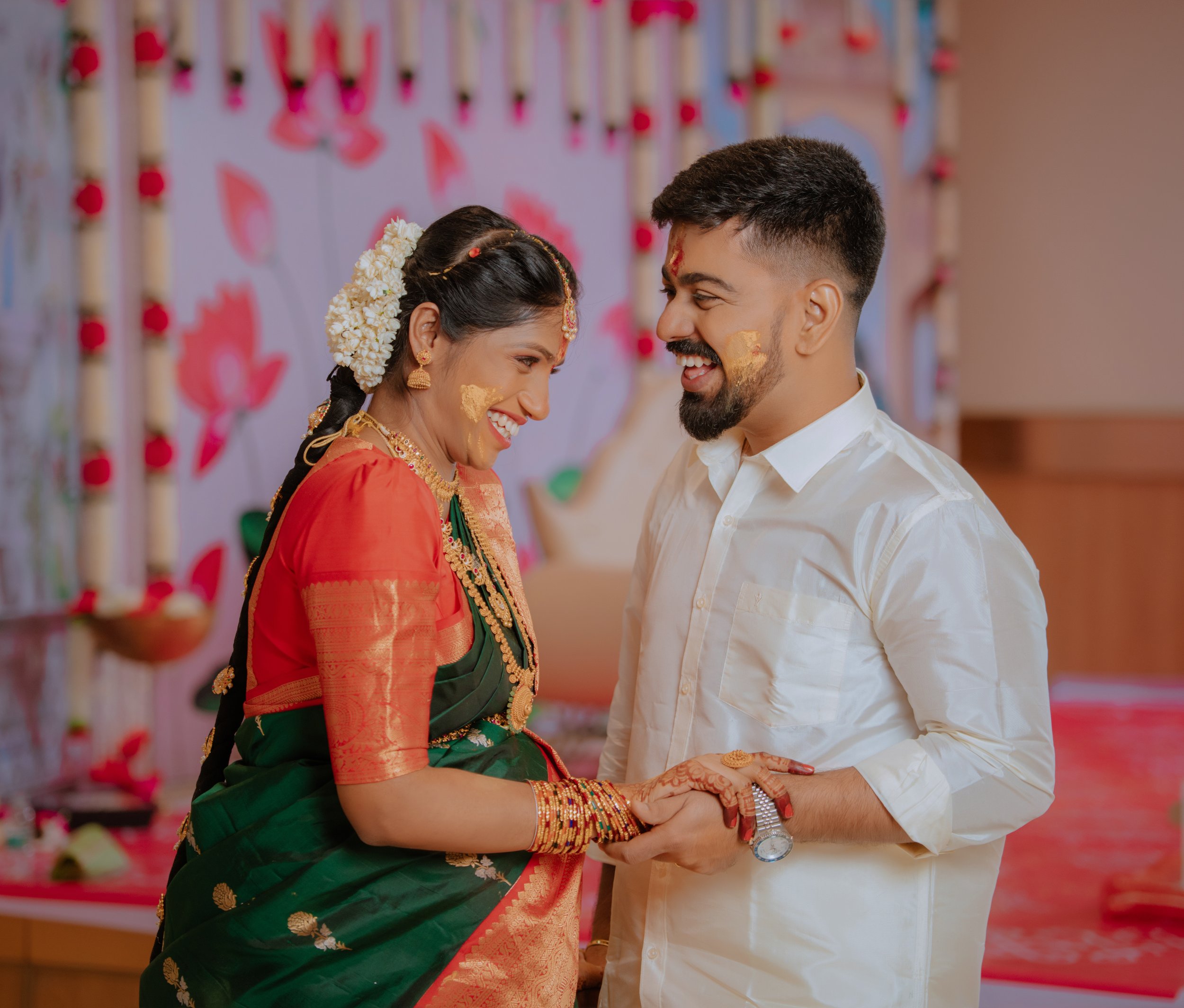 A couple celebrating an Indian wedding, the woman in traditional saree and jewelry, the man in white shirt, both with turmeric marks on their faces, exchanging smiles and holding hands.