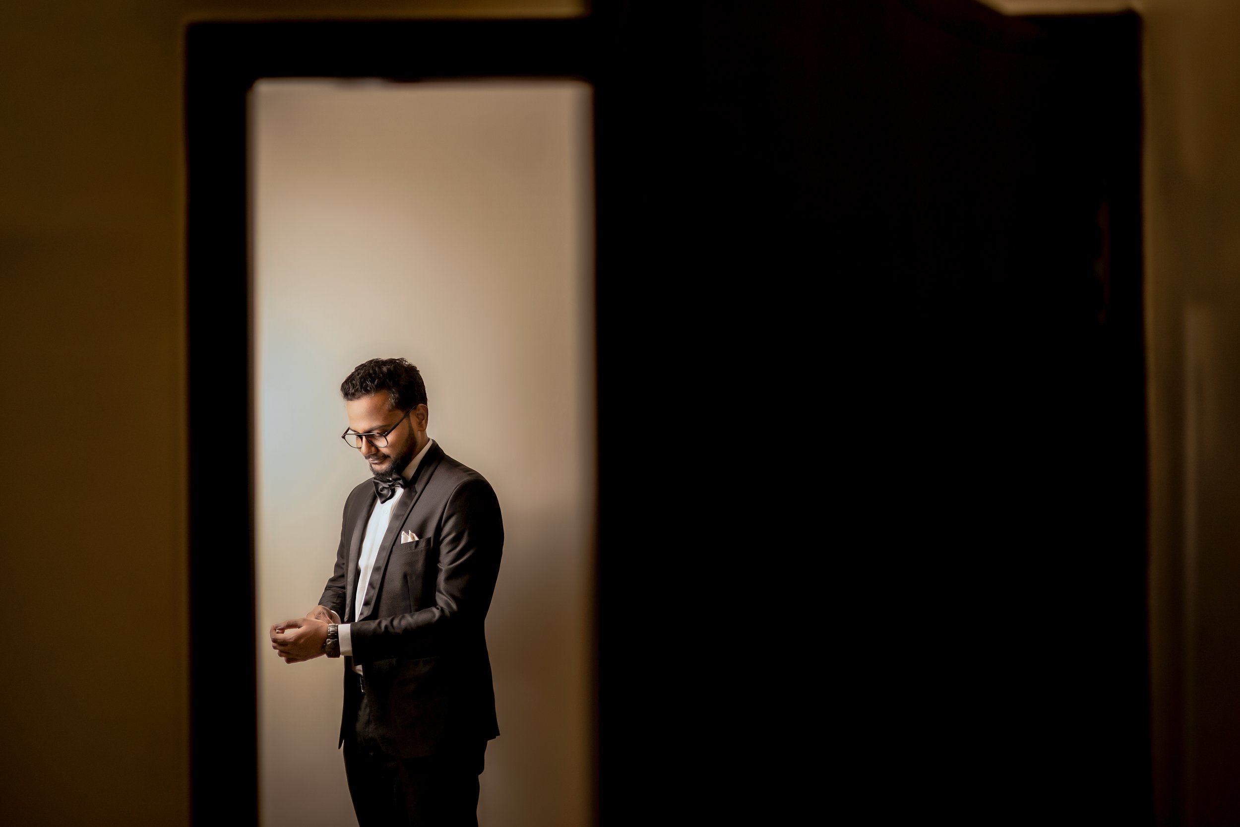 A man dressed in a black tuxedo with a bow tie, glasses, and a watch, standing and looking down at his hands in front of a plain light-colored background, framed by a doorway or opening.