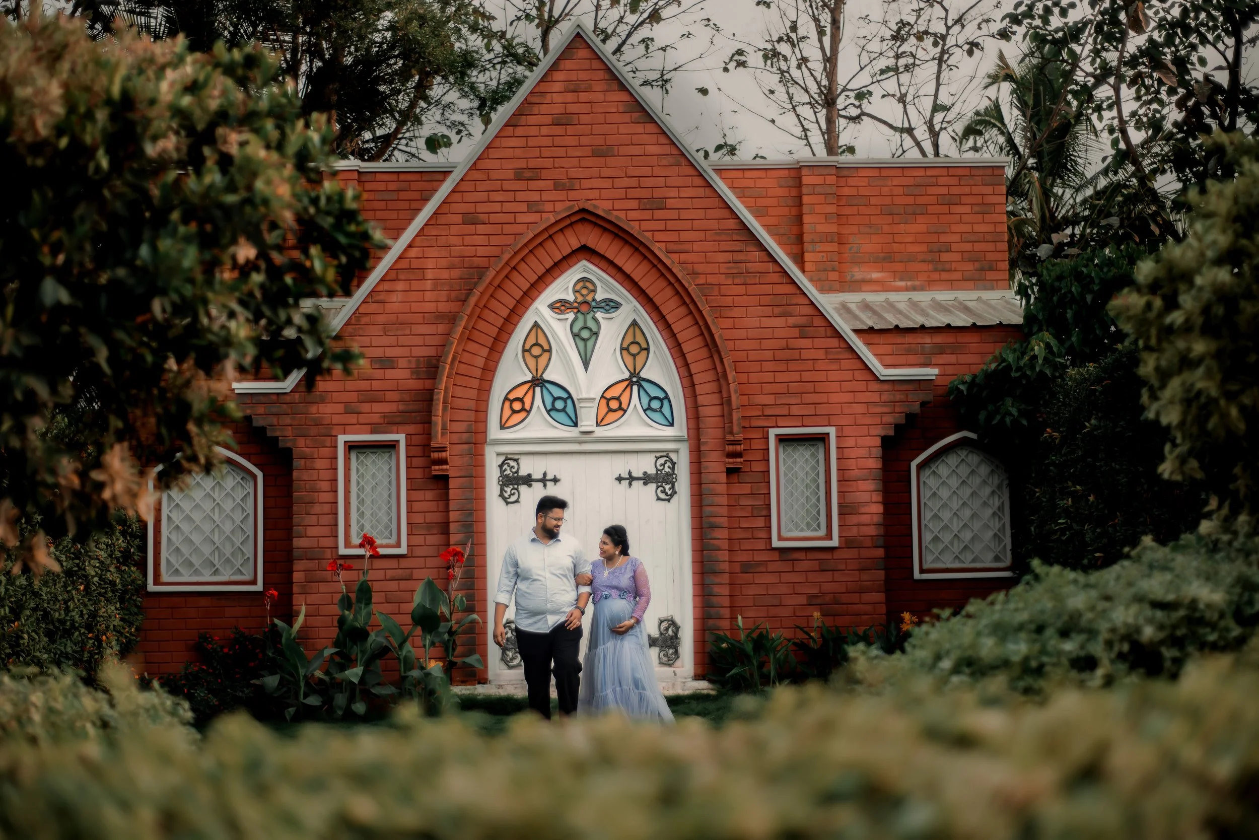 A couple walks hand in hand in front of a small red brick church with stained glass windows and a wooden door, surrounded by greenery and flowers.