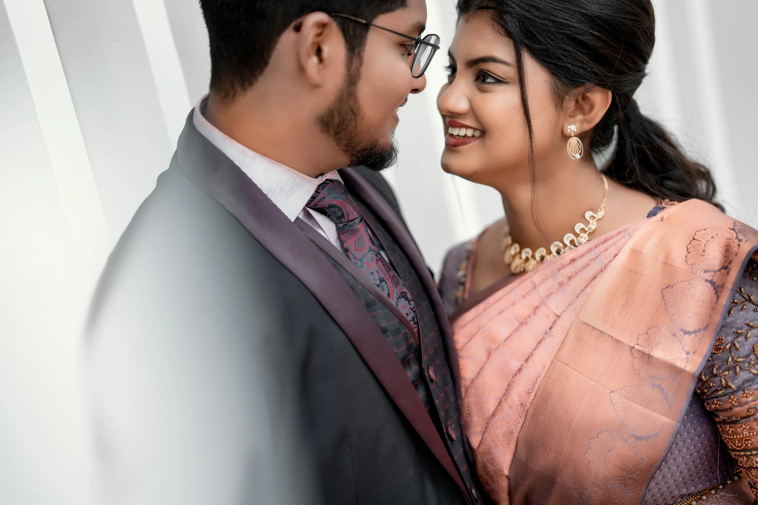 A couple facing each other closely with smiles, standing in front of a white background. The man is wearing a dark suit, checkered shirt, and patterned tie, with glasses. The woman is dressed in traditional Indian attire, including a peach saree with