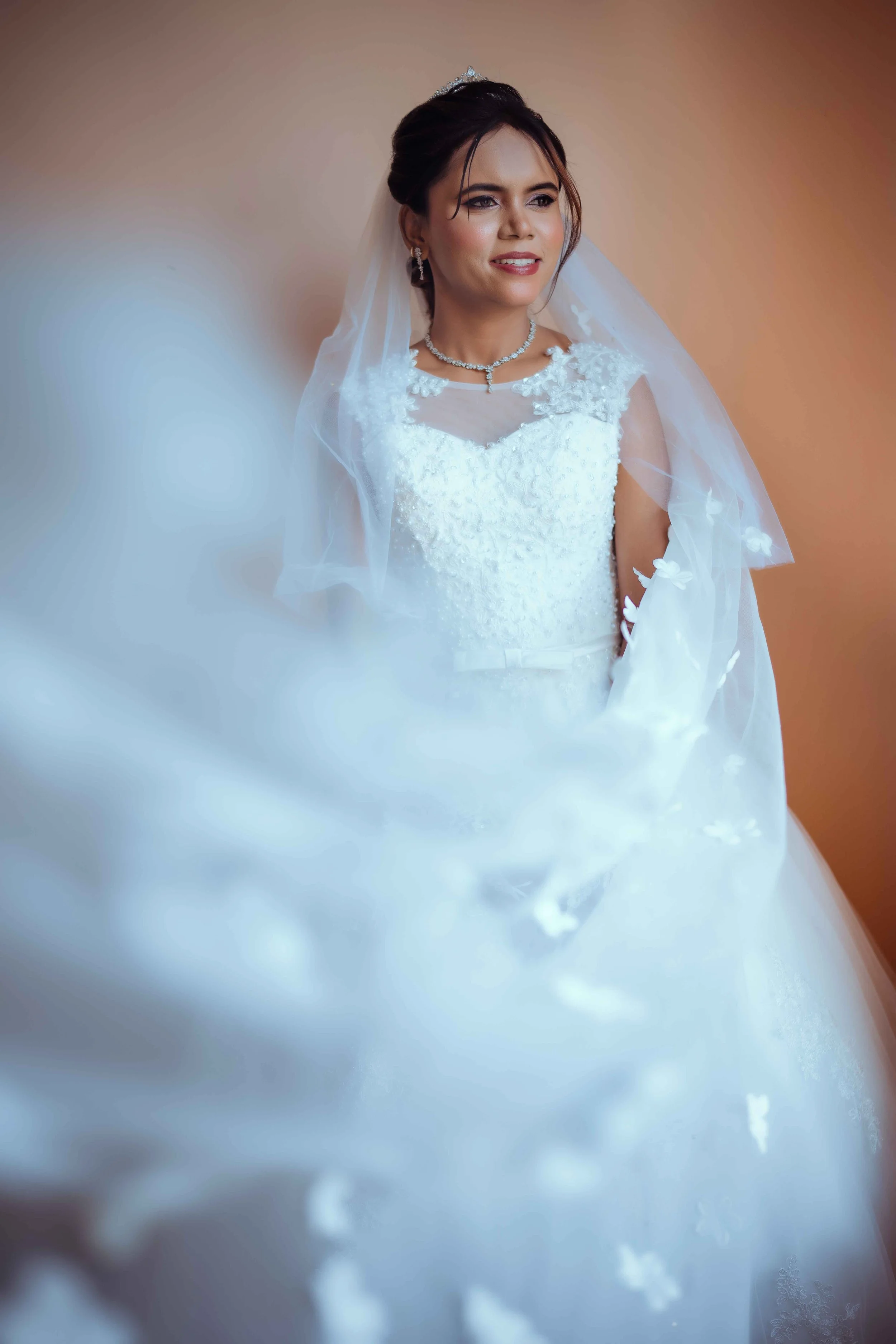 A bride in a white wedding dress with lace details, wearing a necklace and earrings, standing against a beige background.