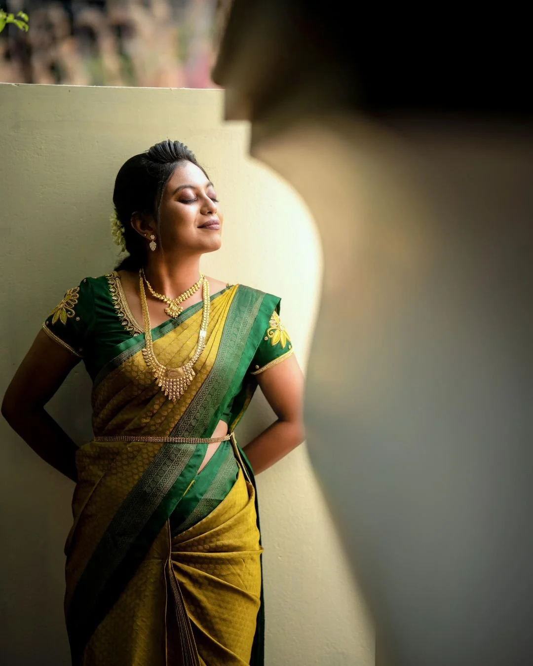 A woman wearing a traditional green and yellow saree with gold jewelry, standing with her eyes closed and smiling softly in front of a light-colored wall.