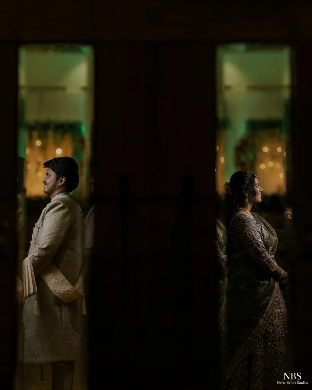A man and woman dressed in traditional Indian attire standing back to back, separated by a dark partition, with windows in the background creating a colorful, blurred bokeh effect.