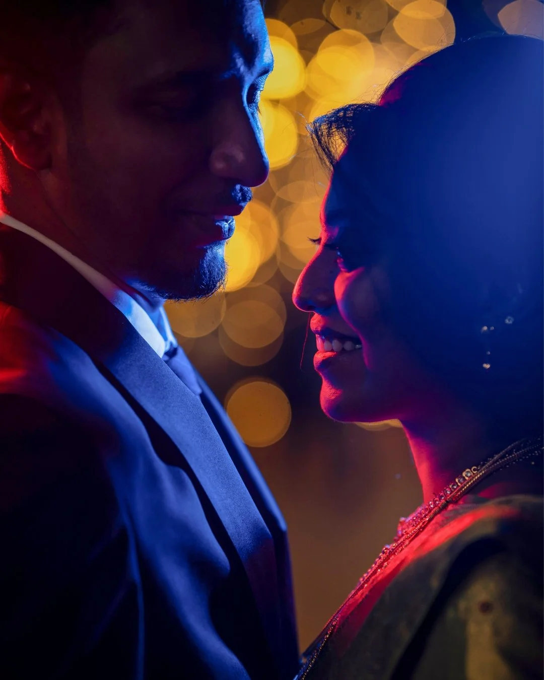 A man and woman face each other close up at night, with warm bokeh lights in the background, smiling and sharing an intimate moment.