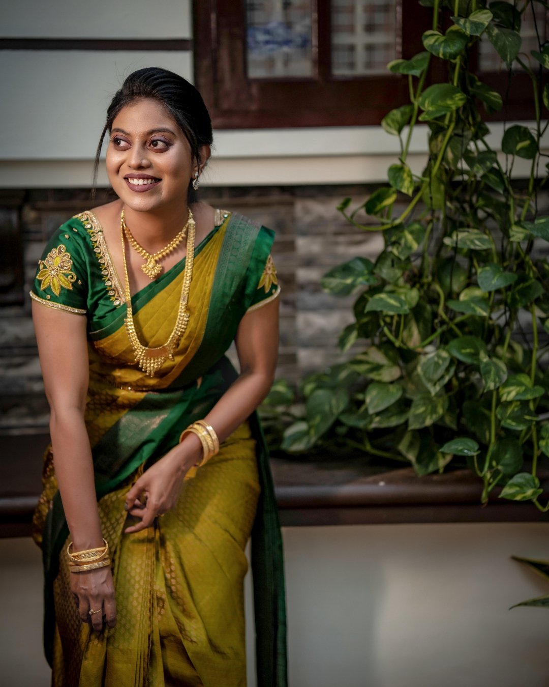 A woman dressed in a traditional green and yellow saree adorned with gold jewelry, smiling and standing near green plants in front of a house.