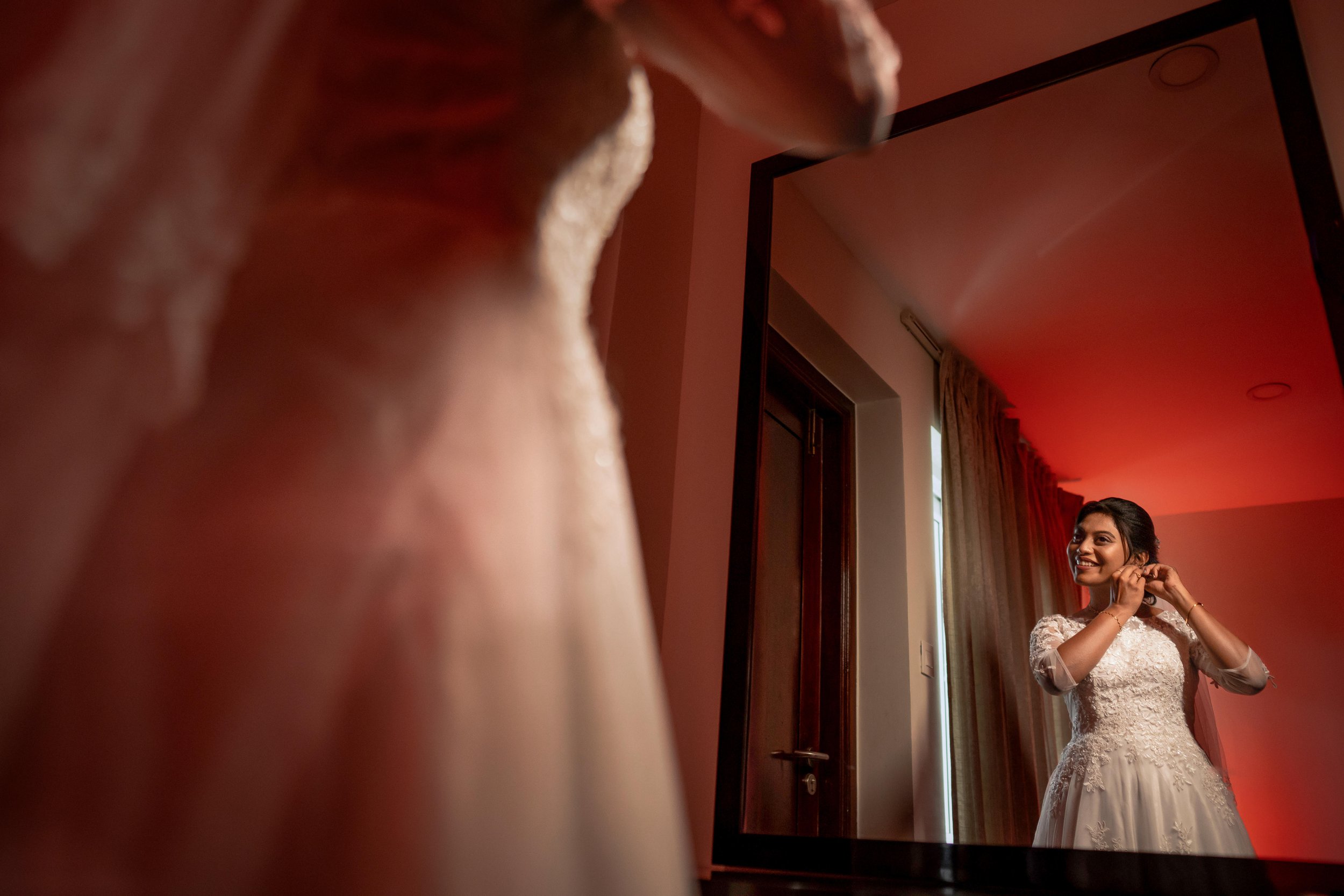 The Bride in a wedding dress smiling and adjusting her earrings while looking at herself in a mirror.