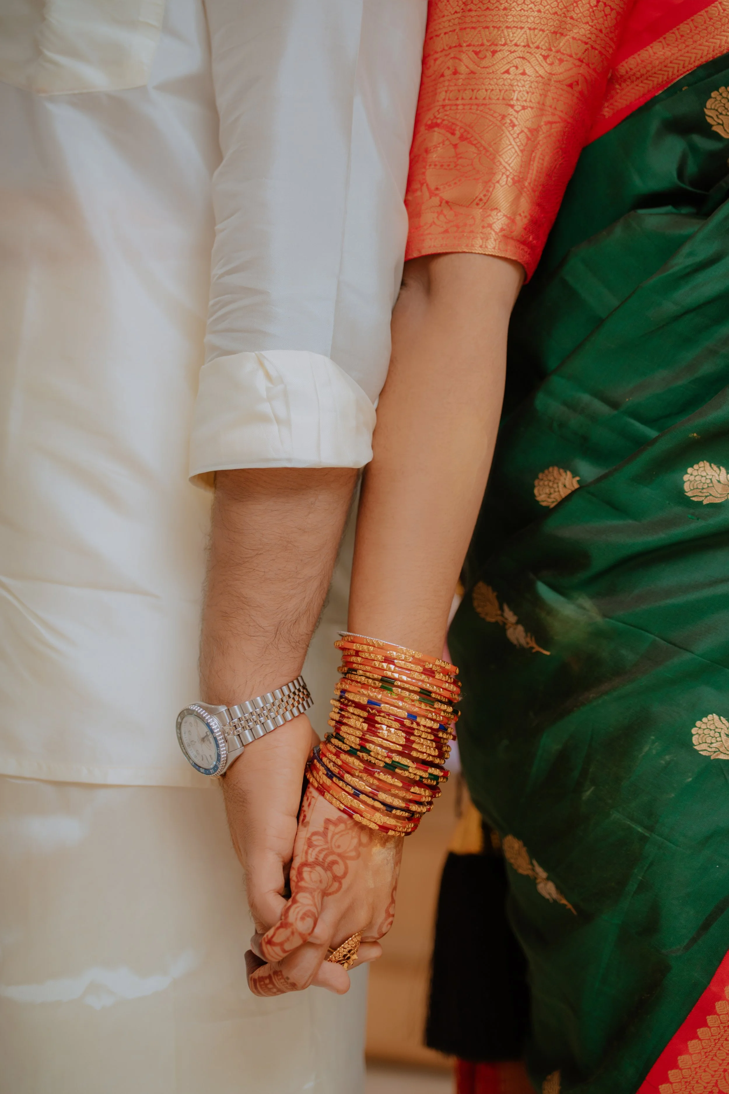 Close-up of a couple holding hands, the man is wearing a watch and a traditional Indian outfit, the woman has henna on her hand and is wearing multiple red and gold bangles, with green and red traditional attire.