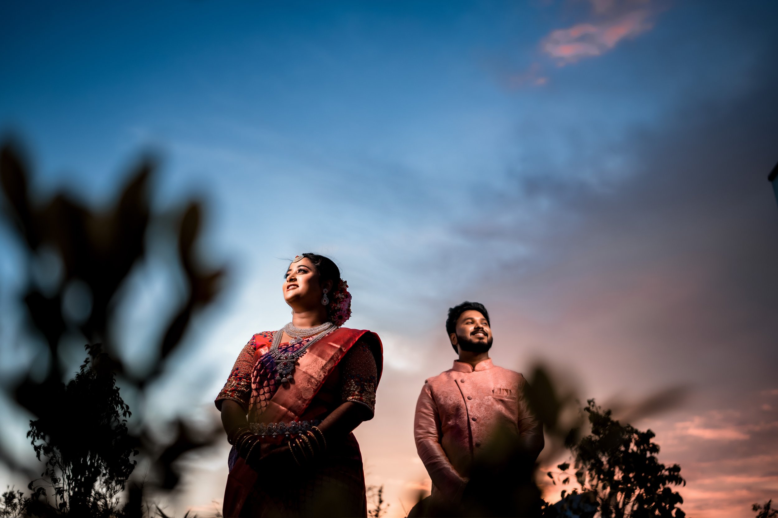 The Bride and groom dressed in traditional Indian attire standing outdoors during sunset, smiling and looking towards the sky.