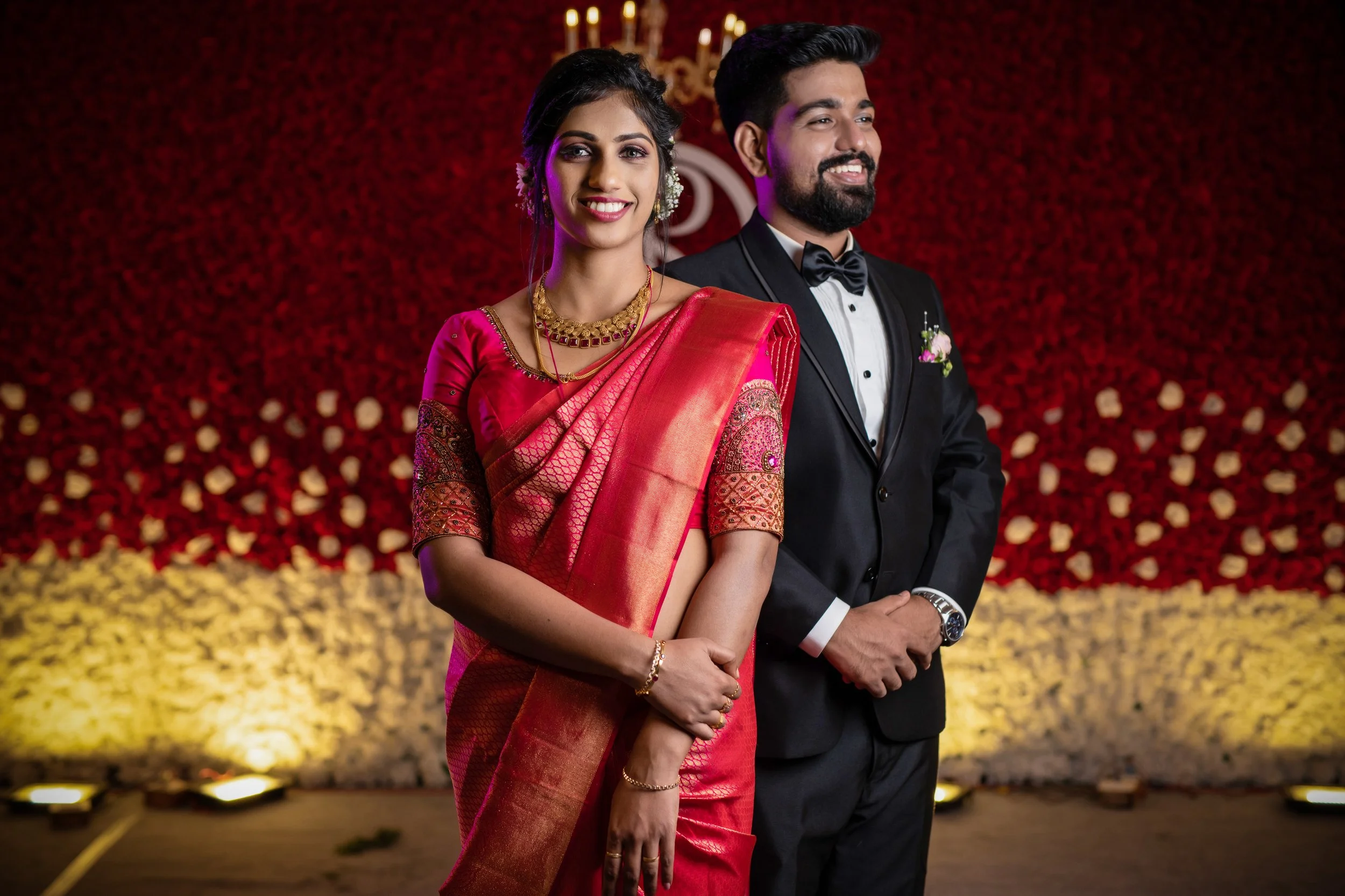 A smiling Indian woman in a red traditional saree with gold jewelry standing next to a man in a black tuxedo at a celebration event with a red and yellow floral backdrop.