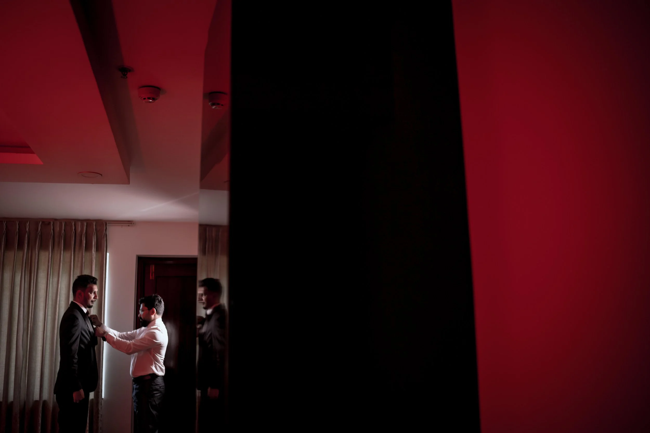 Men in suits, one adjusting another's tie, in a dimly lit room with a red and black color scheme.