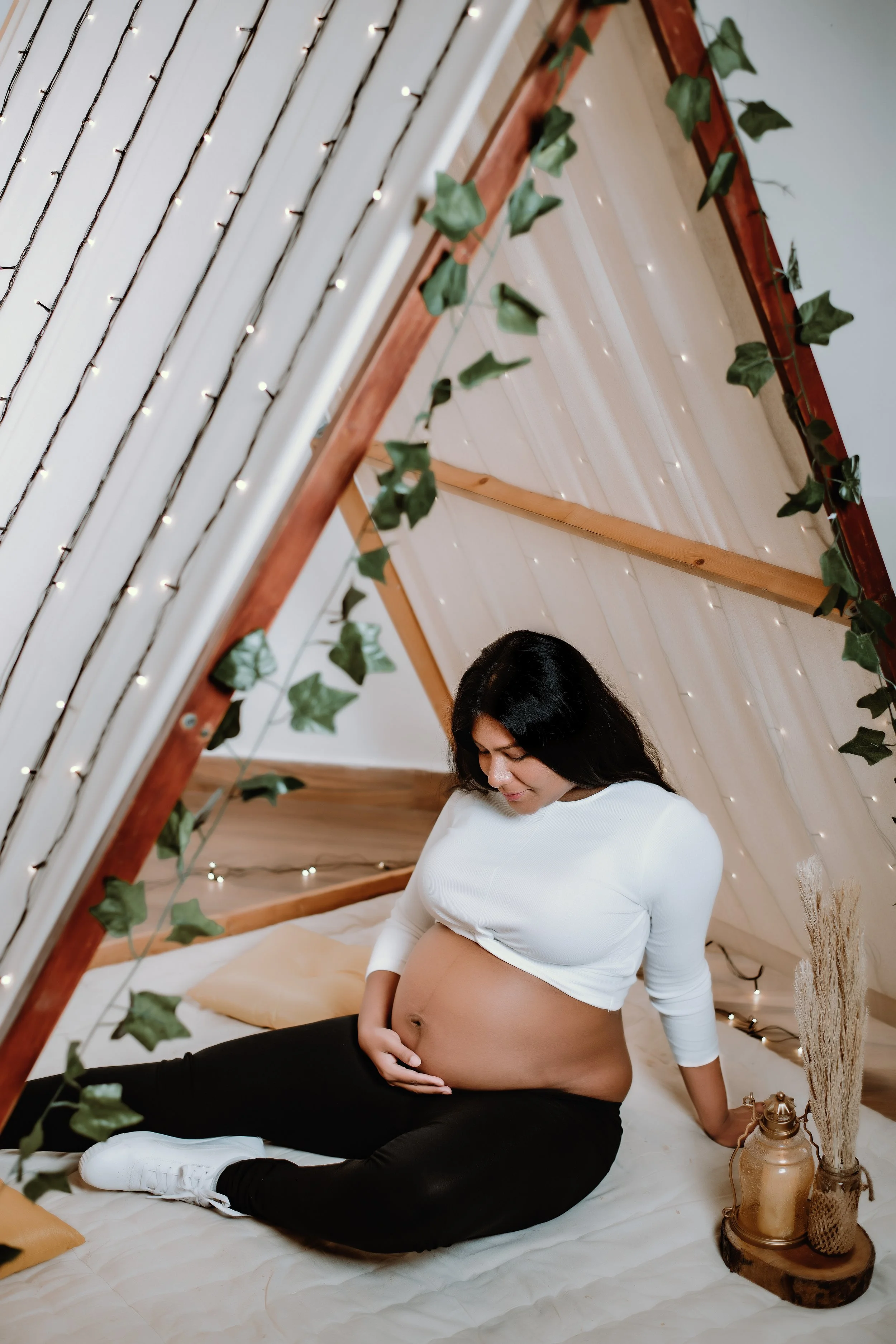 The Bride - Soon to be mother - sitting inside an A-frame tent decorated with string lights and ivy, touching her belly gently.