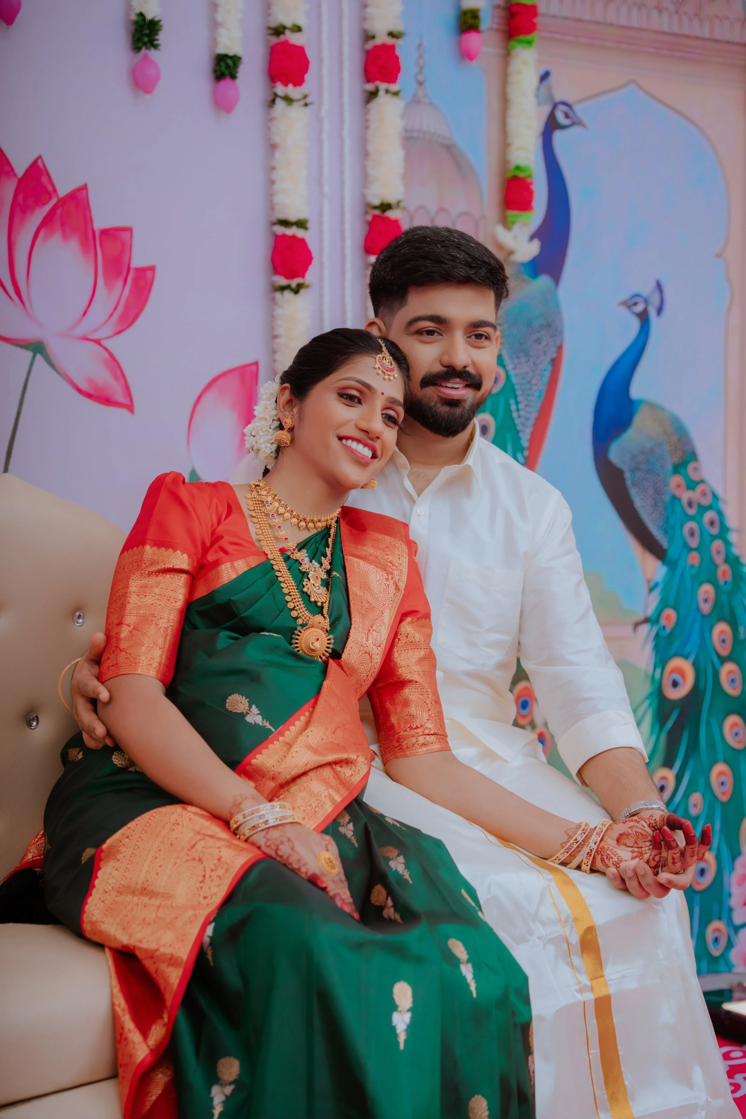 A couple dressed in traditional Indian wedding attire sitting together, smiling. The woman wears a green and red saree with gold jewelry and flowers in her hair. The man wears a white dhoti and kurta. The background features colorful peacock and lotu