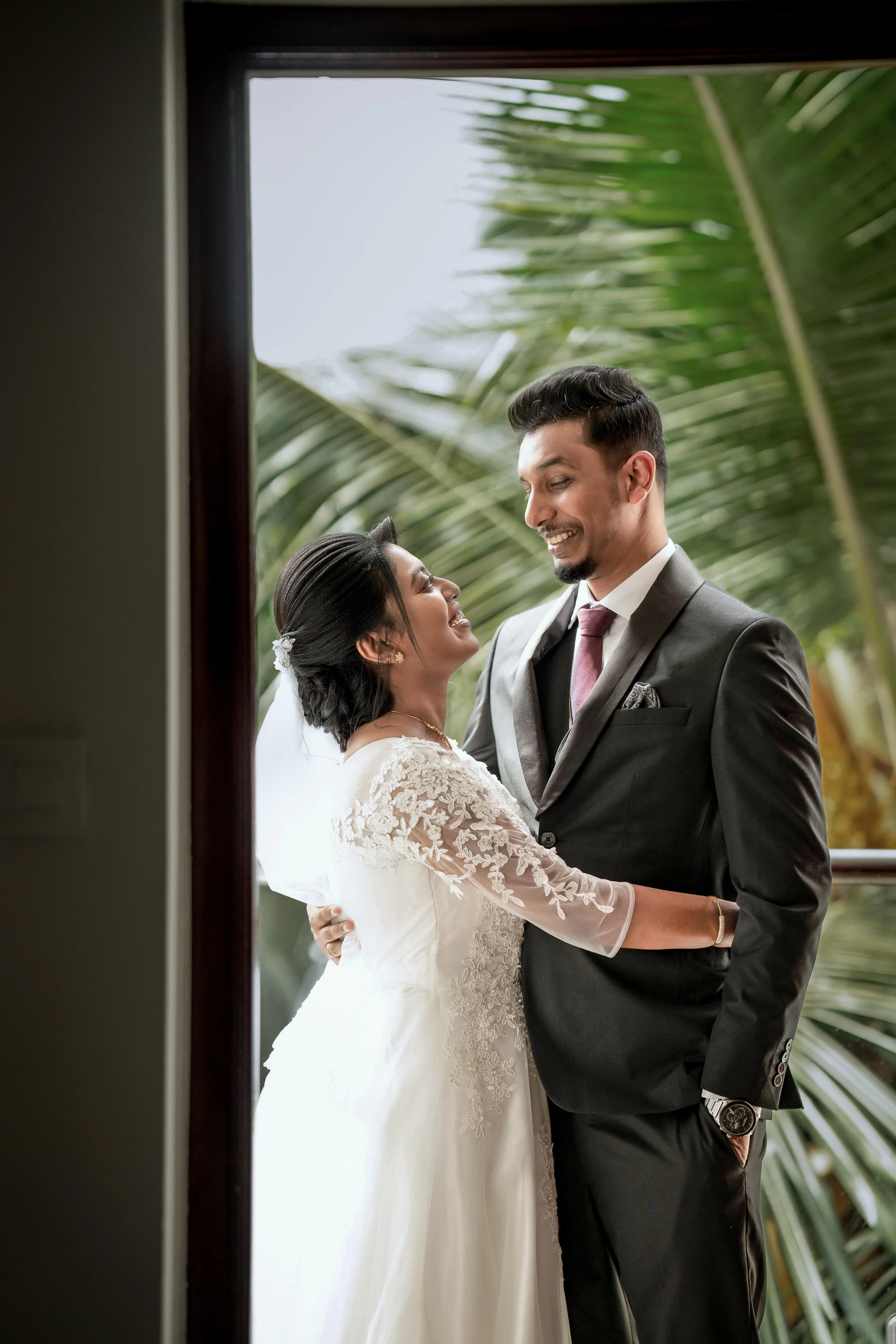 A bride and groom in wedding attire sharing a happy moment outside with tropical palm trees in the background.