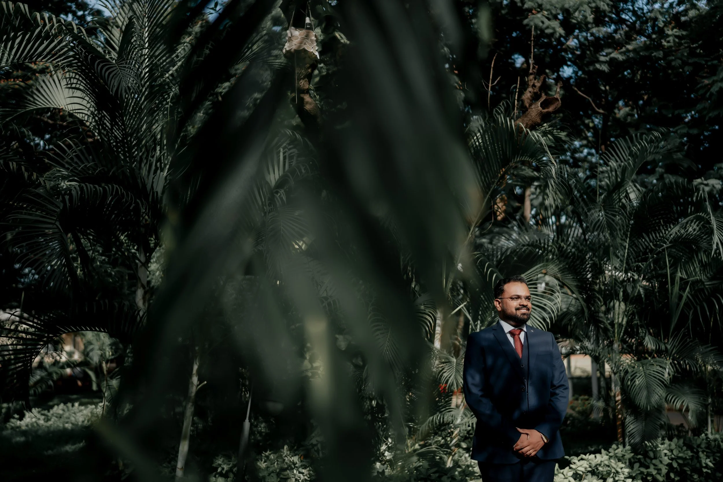 The Bride in a dark suit, red tie, and glasses standing outdoors among lush green tropical plants, smiling with his hands folded in front of him.