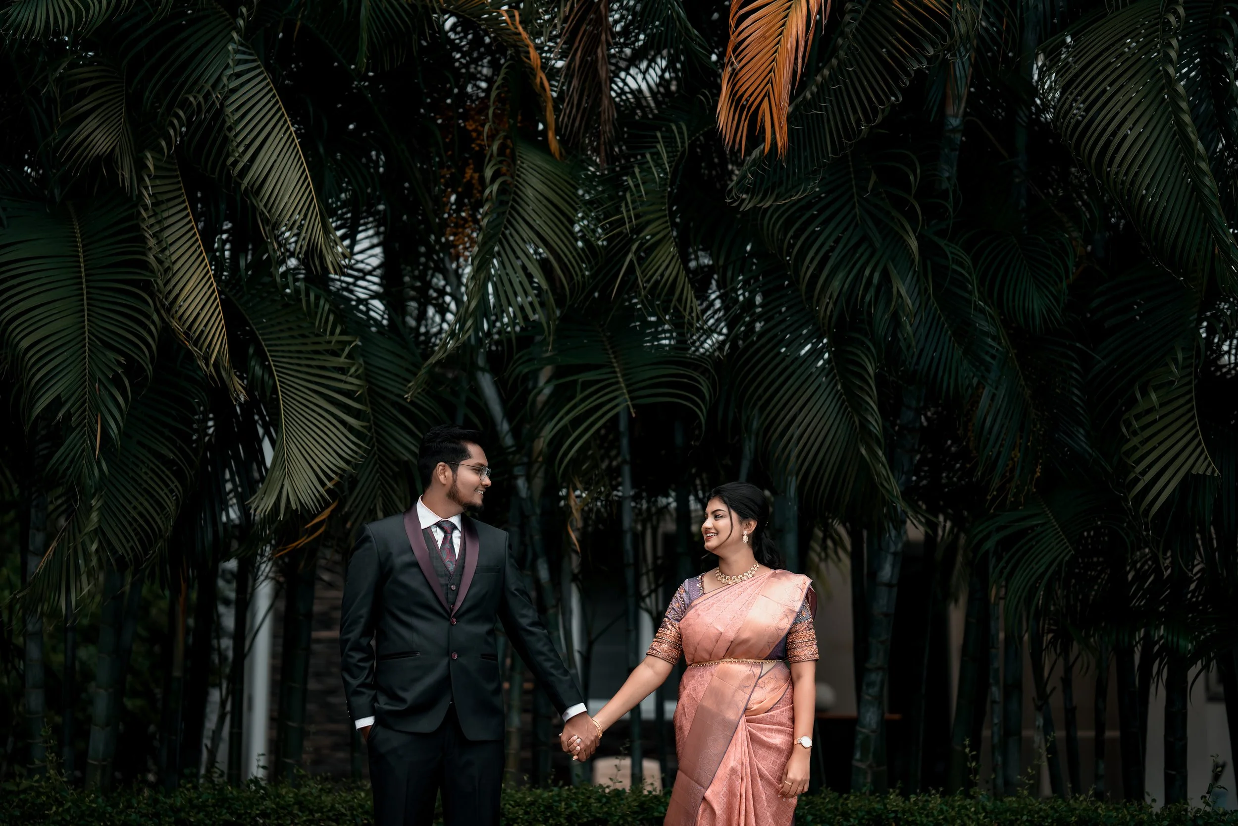 Bride and groom holding hands outdoors, dressed in formal attire, with large palm leaves in the background.