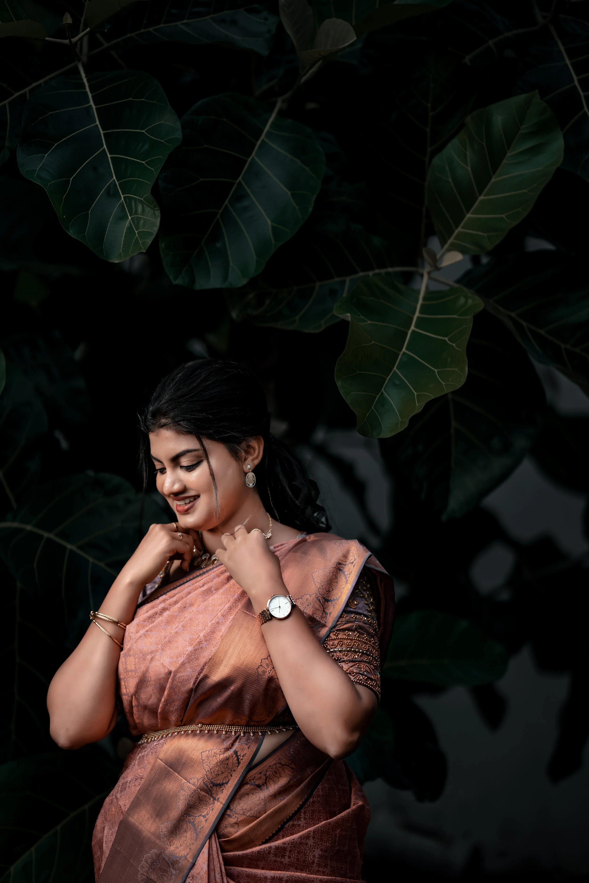The Bride in a traditional saree standing amidst large green leaves, looking down with a gentle smile.