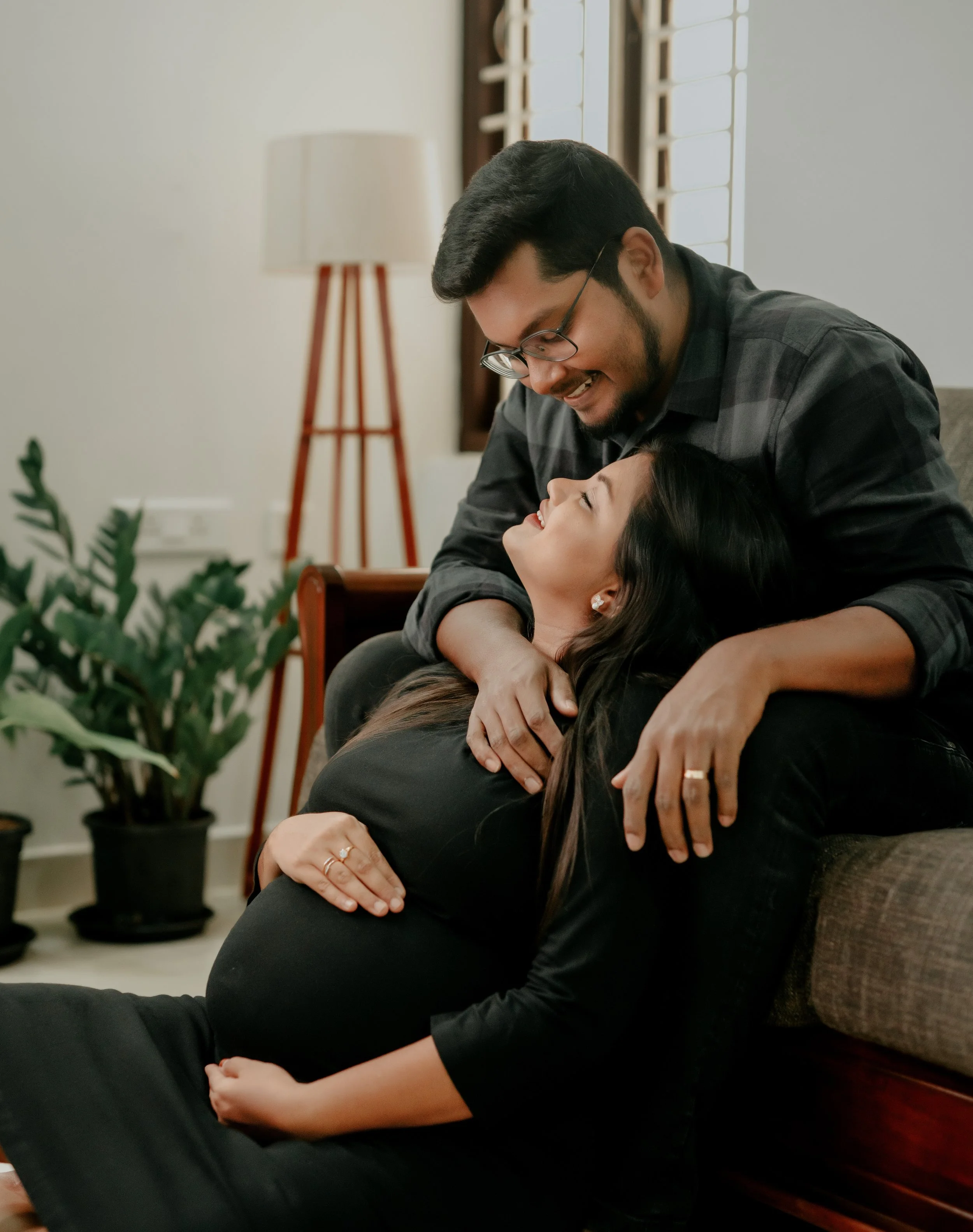A couple celebrating pregnancy at home, with the woman sitting on the floor and the man leaning over her, both smiling and looking happy.