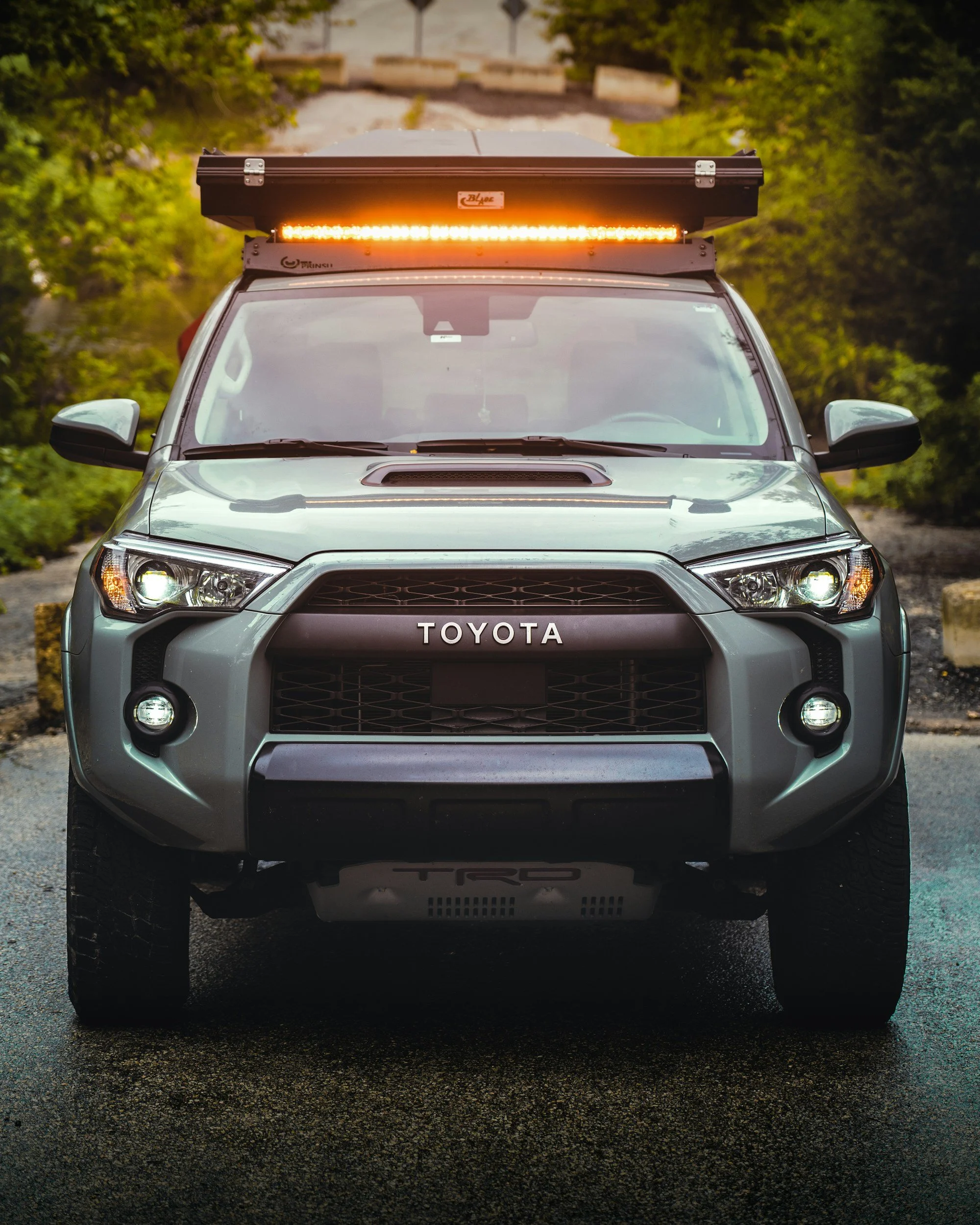 Front view of a silver Toyota off-road vehicle with an LED light bar mounted on the roof, parked on a wet asphalt road with a background of trees and bushes.