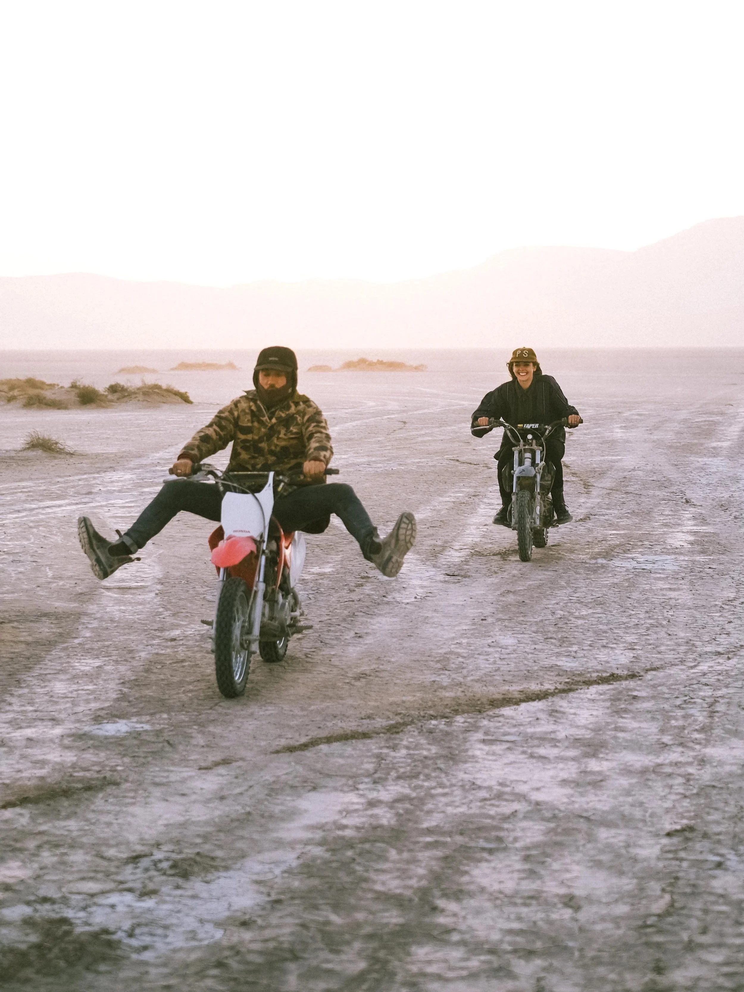 Two people riding bikes on a dry, cracked desert landscape with mountains in the background, one person has their legs extended outward while riding.