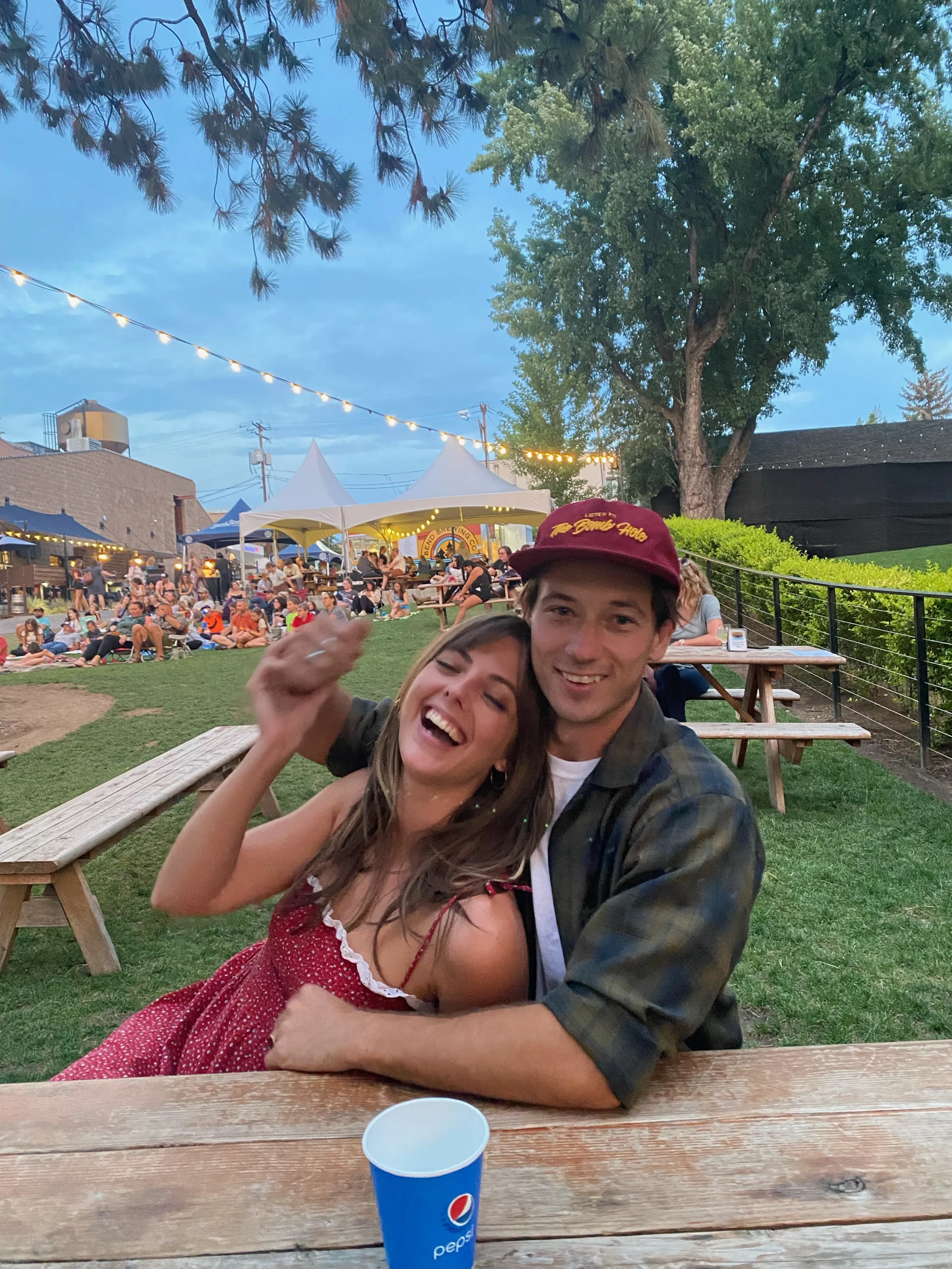 A happy couple sitting at a picnic table outdoors at a lively event during the evening. The woman is laughing and leaning into the man, who is smiling. There are many people in the background under string lights, with tents and trees around.