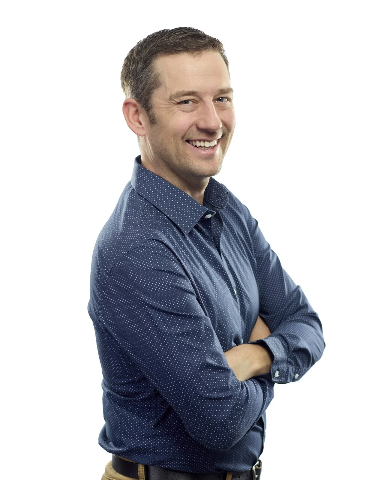 A smiling man with short brown hair, wearing a blue polka dot shirt, standing with arms crossed against a white background.