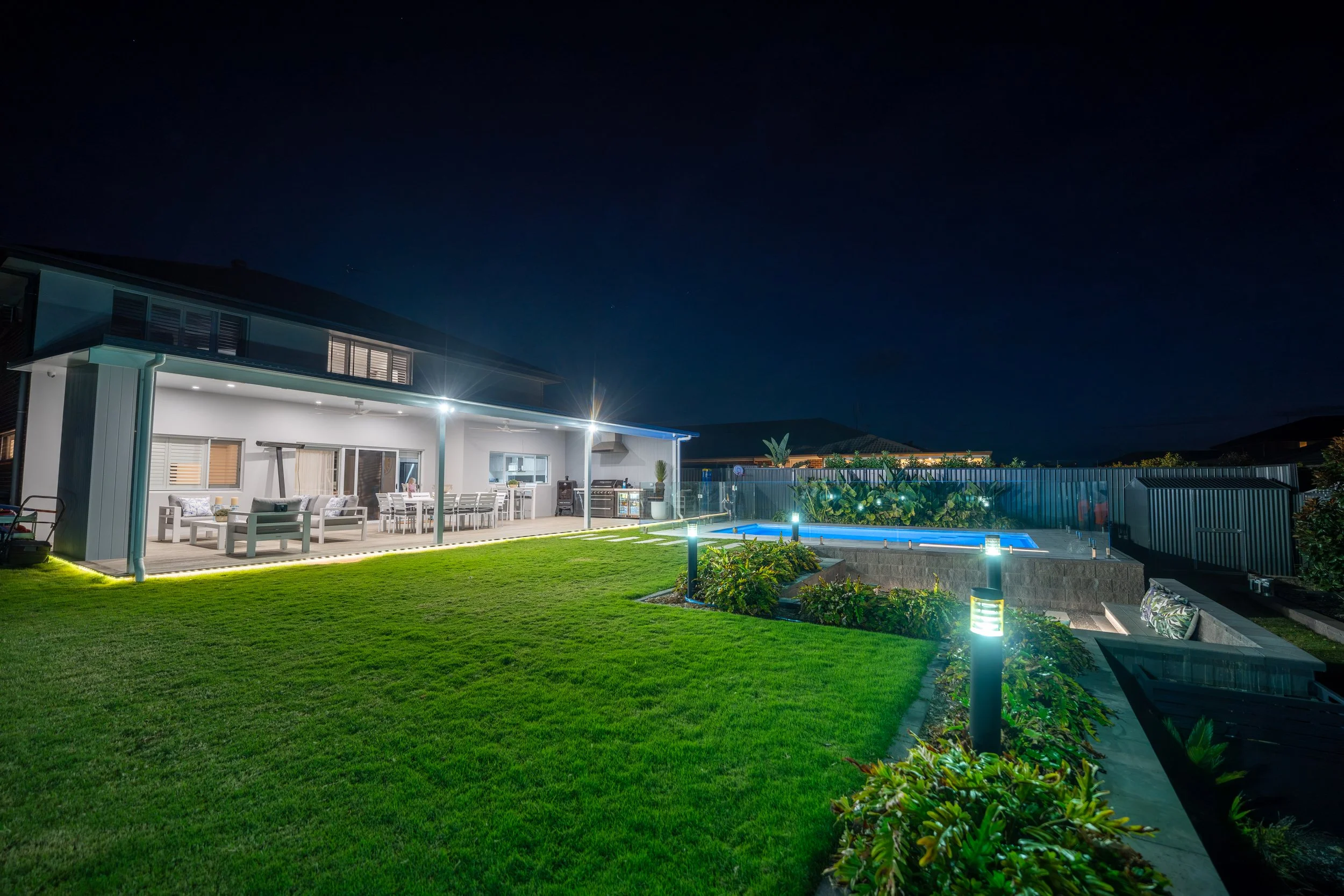 Nighttime view of a modern house with a backyard patio, illuminated with outdoor lights, a swimming pool, and a well-maintained lawn, under a dark sky.