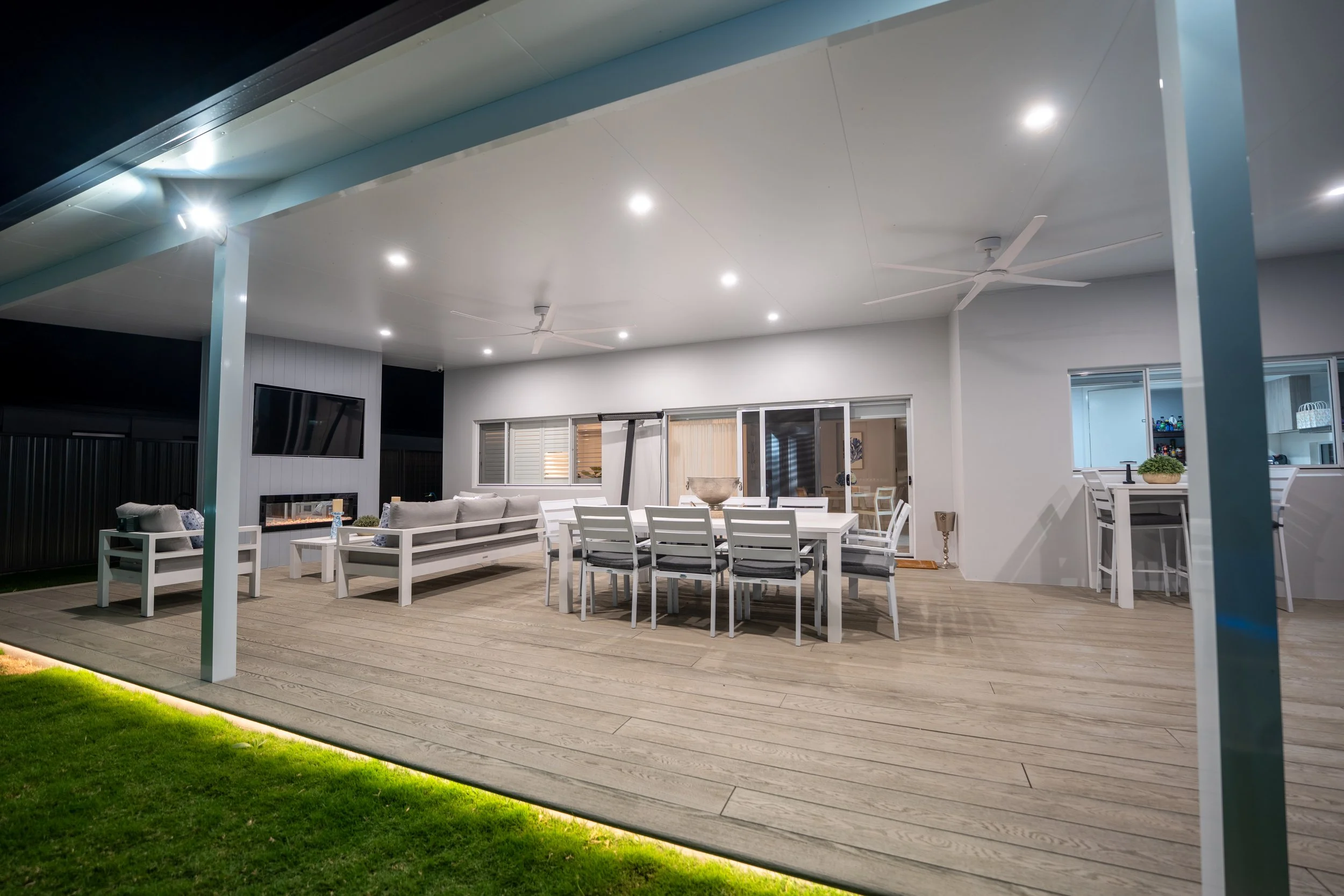 Outdoor covered patio area with white dining table and chairs, ceiling fans, and a fireplace, illuminated at night with lighting and a view of a grassy lawn.