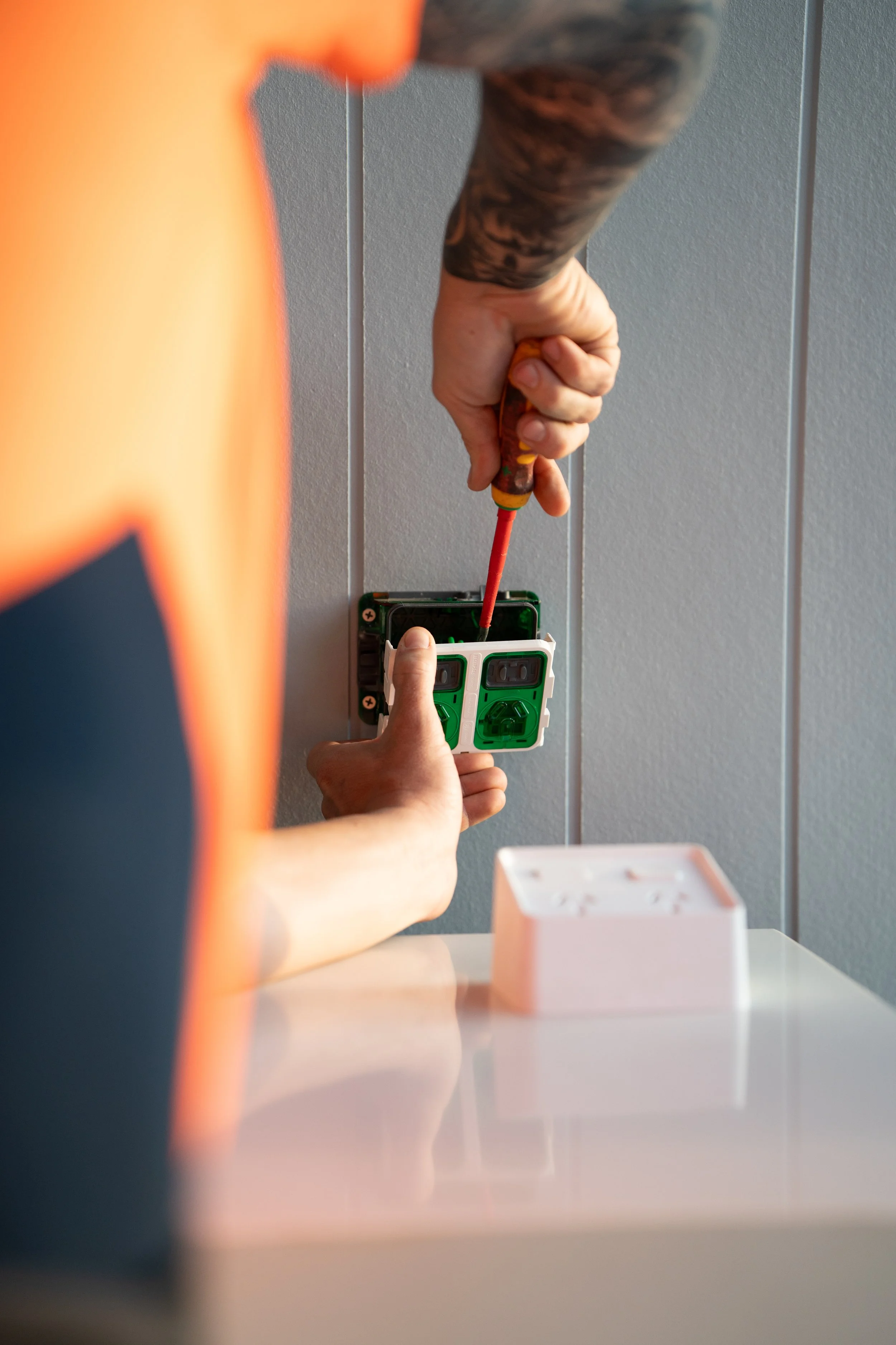 A person using a screwdriver to work on an electrical outlet on a gray wall. The person's left arm is visible, covered by a black marbled shirt, and they are wearing orange gloves.