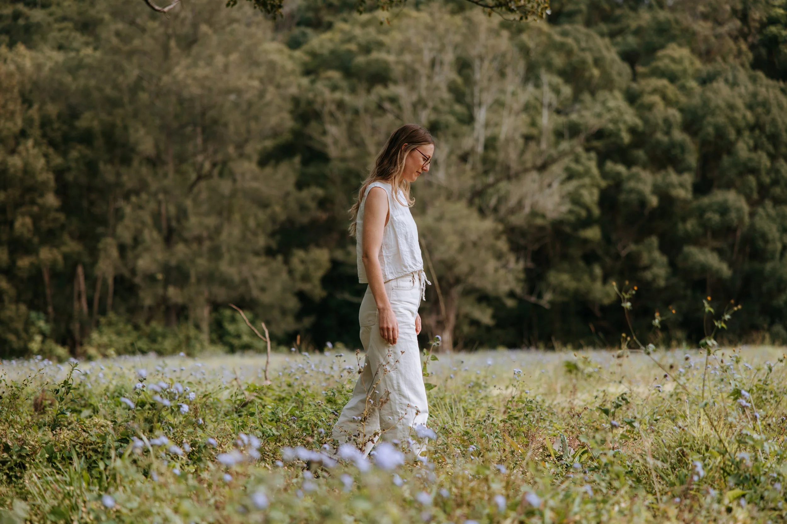 A woman standing alone in a field of wildflowers with trees and forest in the background, wearing a white sleeveless top and beige pants, looking down thoughtfully.