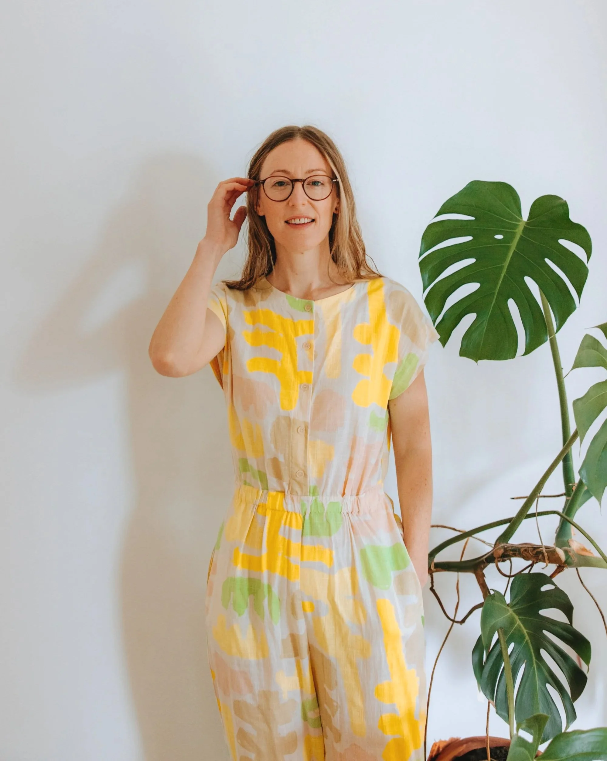 A woman with glasses and long hair, wearing a colorful patterned jumpsuit, standing next to a potted monstera plant against a plain white background.