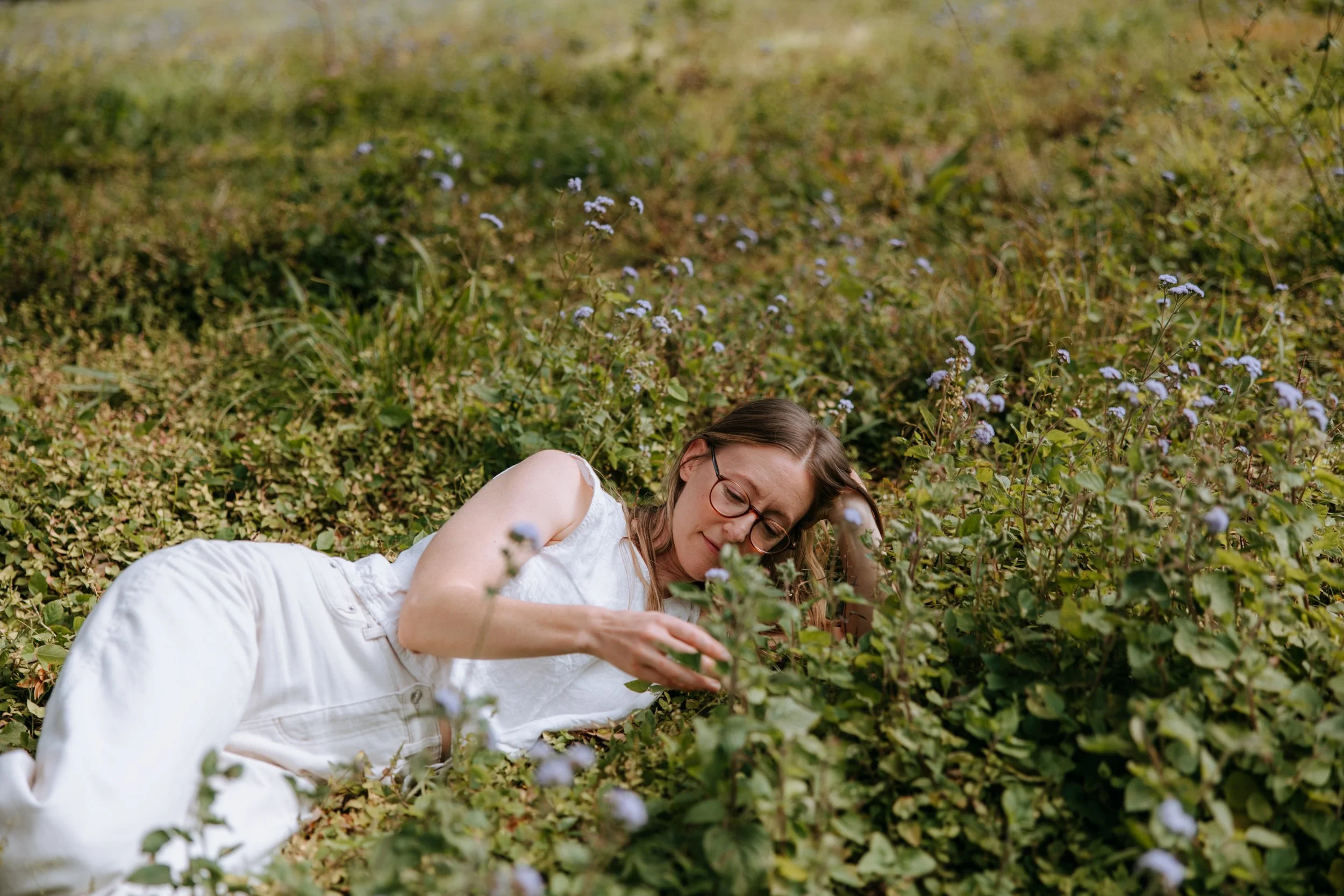 Women lying on the ground in a grassy field with purple flowers, wearing white clothing and glasses.