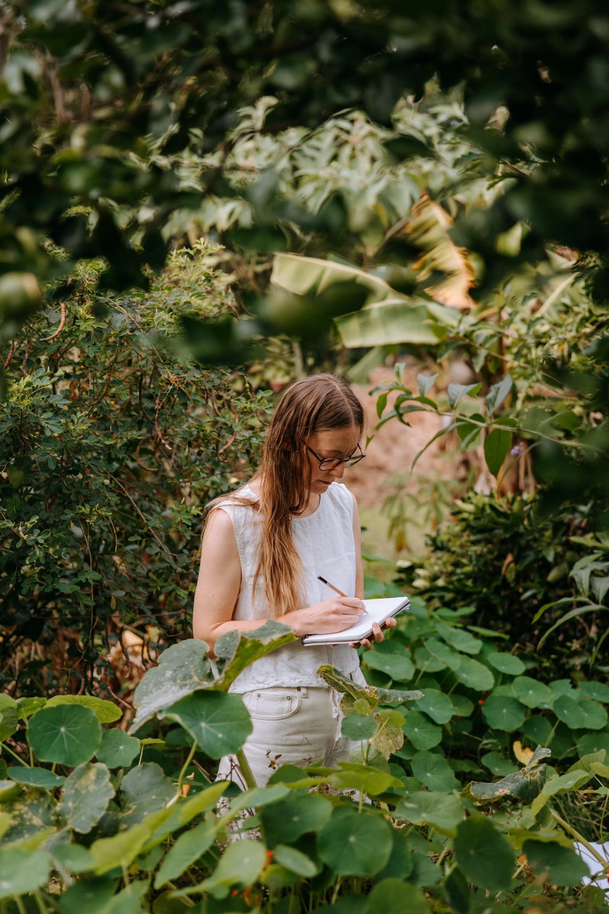 A woman with long hair, glasses, and wearing a sleeveless white top, stands among green leafy plants, writing in a notebook.