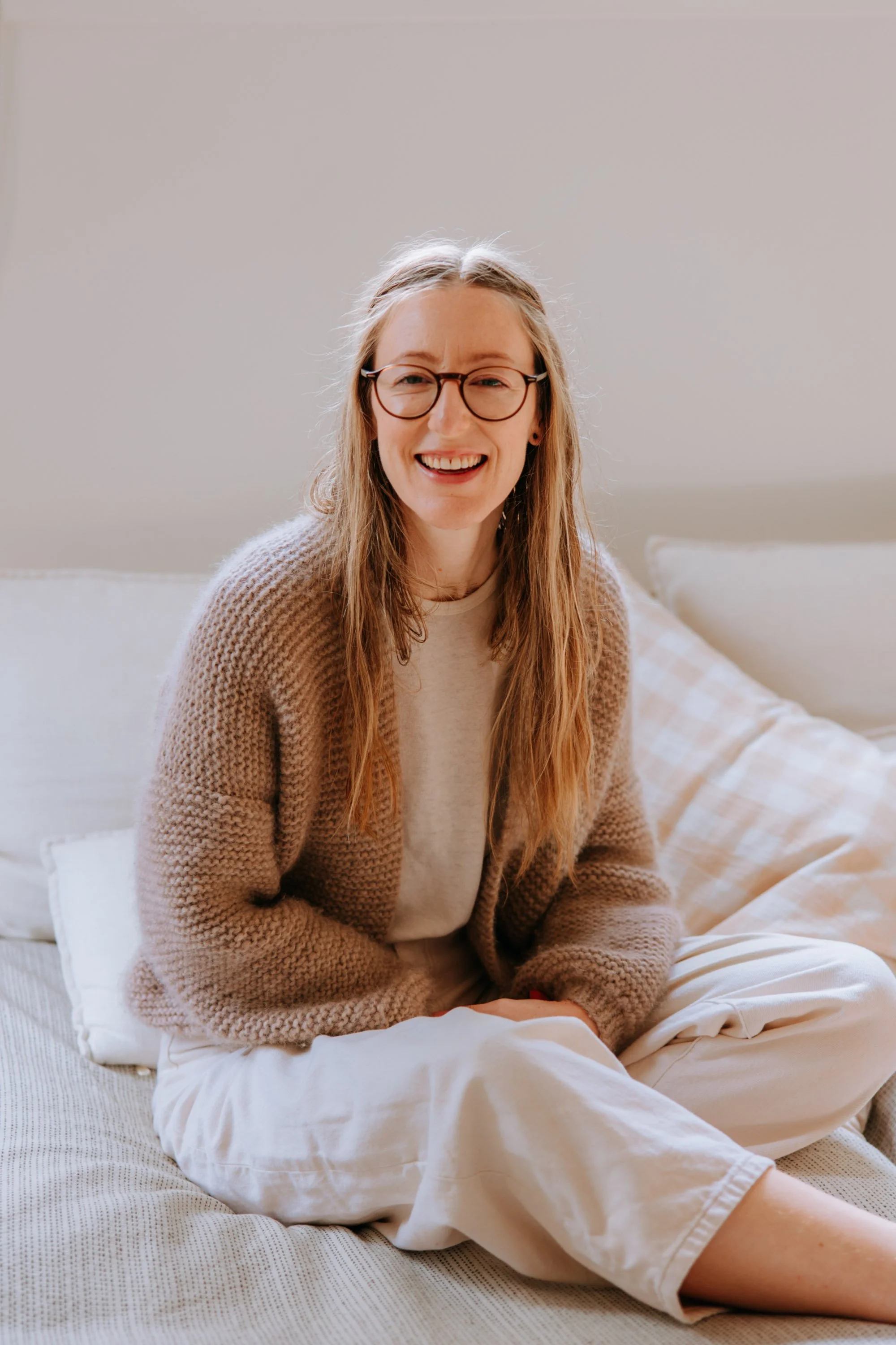 A woman with long blonde hair, glasses, and a brown knitted cardigan sitting on a bed with beige and white bedding, smiling at the camera.