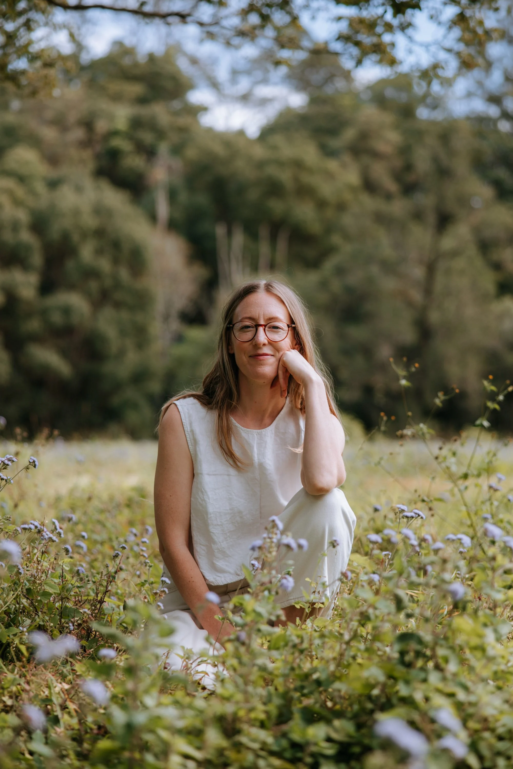 A woman sitting in a field of purple flowers with a background of green trees and a cloudy sky.